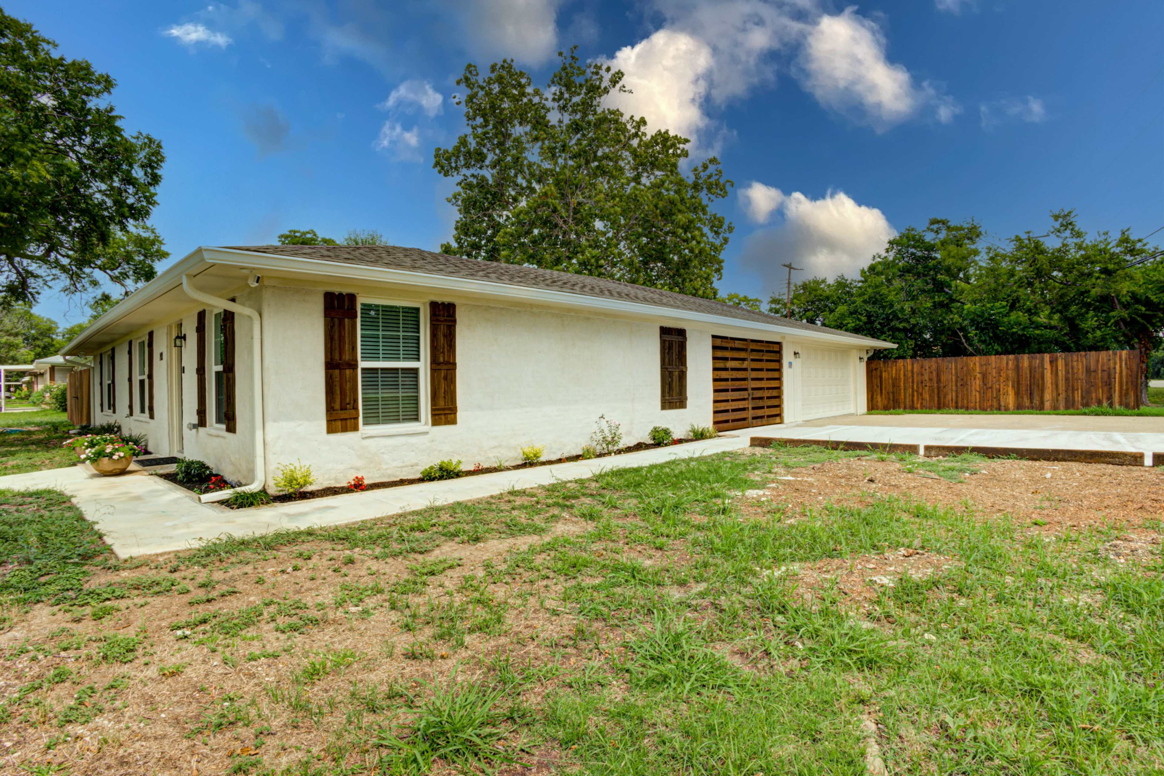 A one-story white house with brown shutters and a garage, surrounded by a grassy yard and a fence in the background.