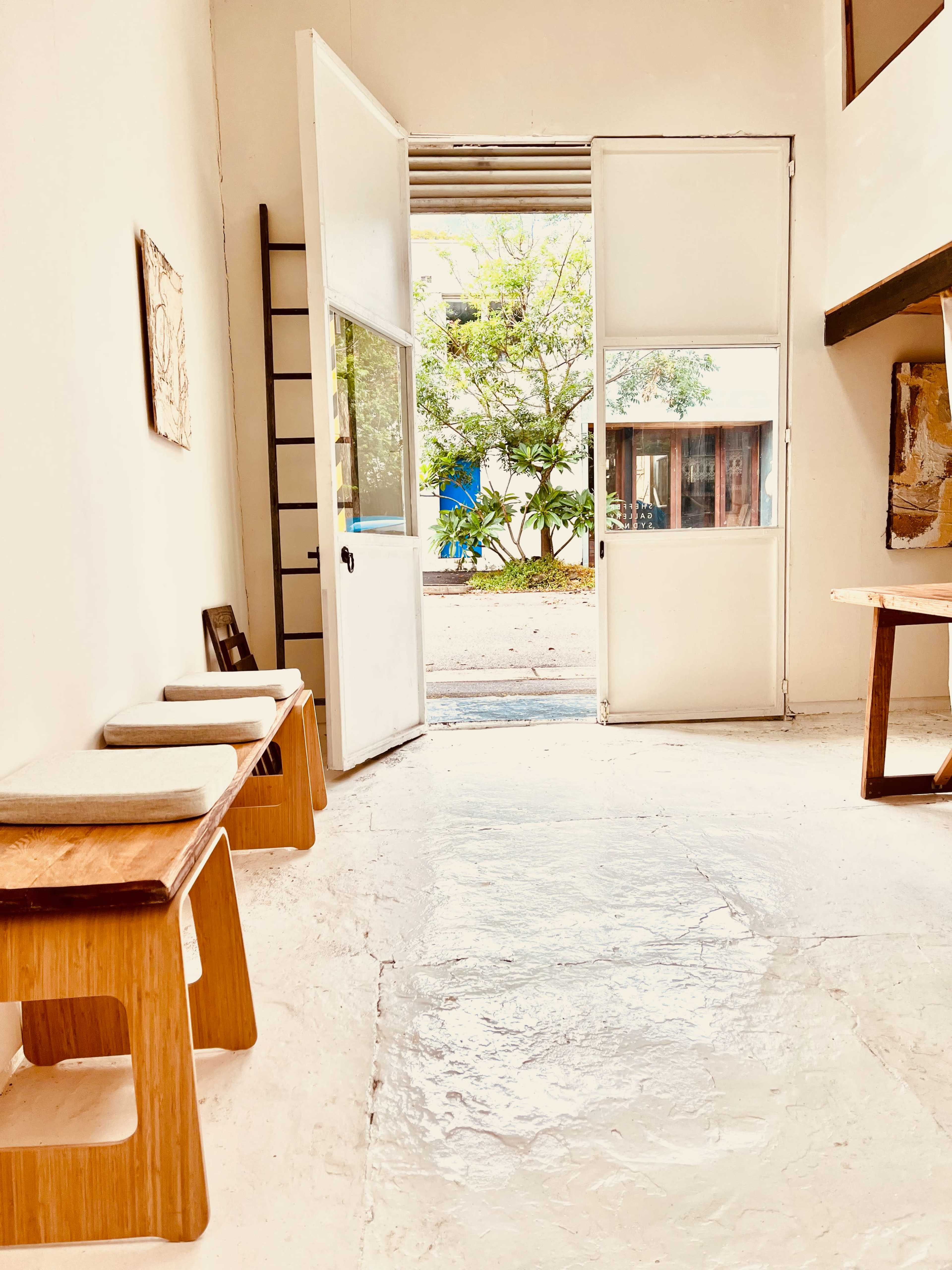 A bright, airy room features an open door leading to a garden, with wooden benches along one wall and a table against another.