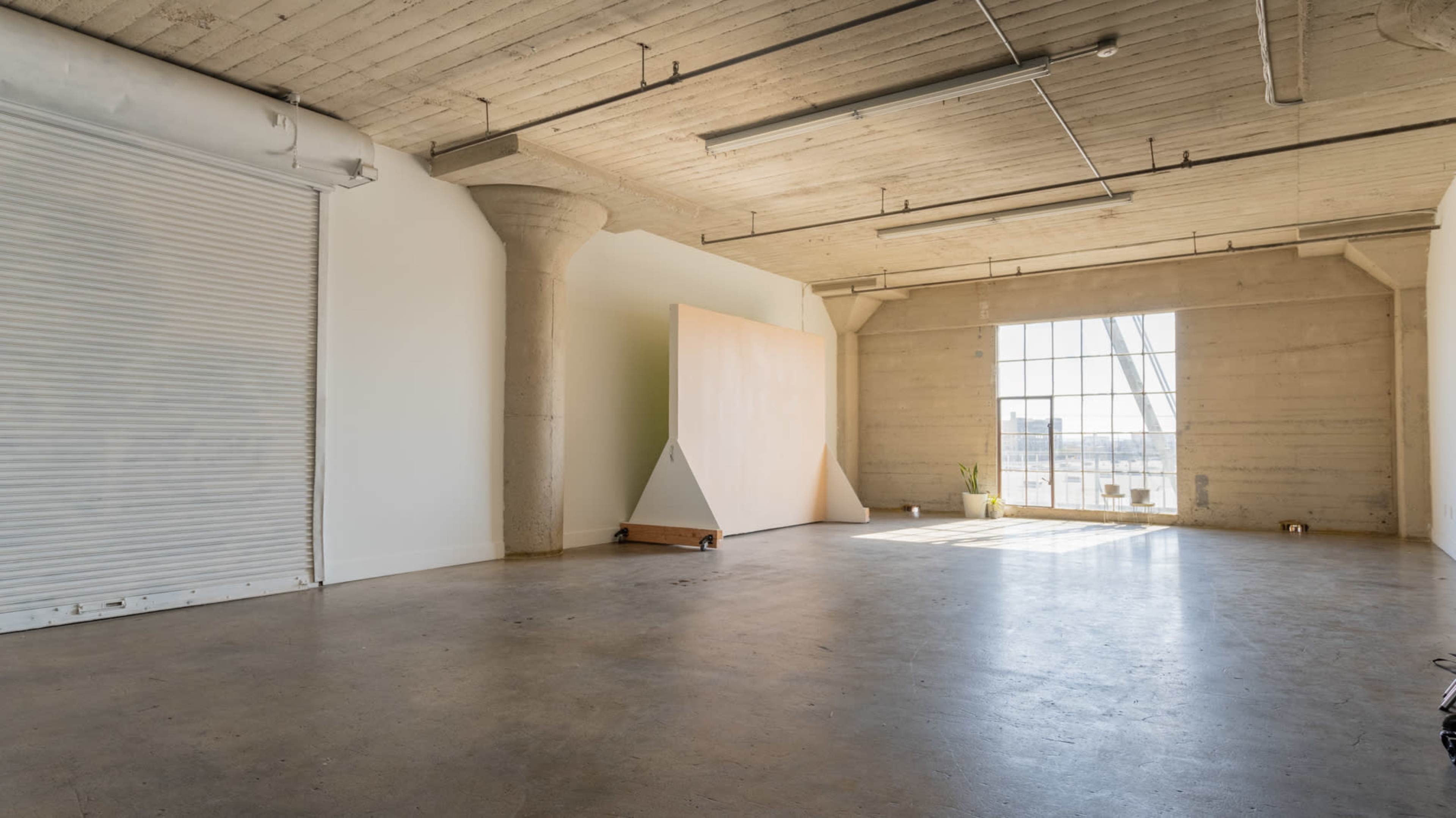 A spacious, empty room with concrete walls, high ceilings, and a large window letting in natural light, featuring a large, triangular wooden panel against one wall.