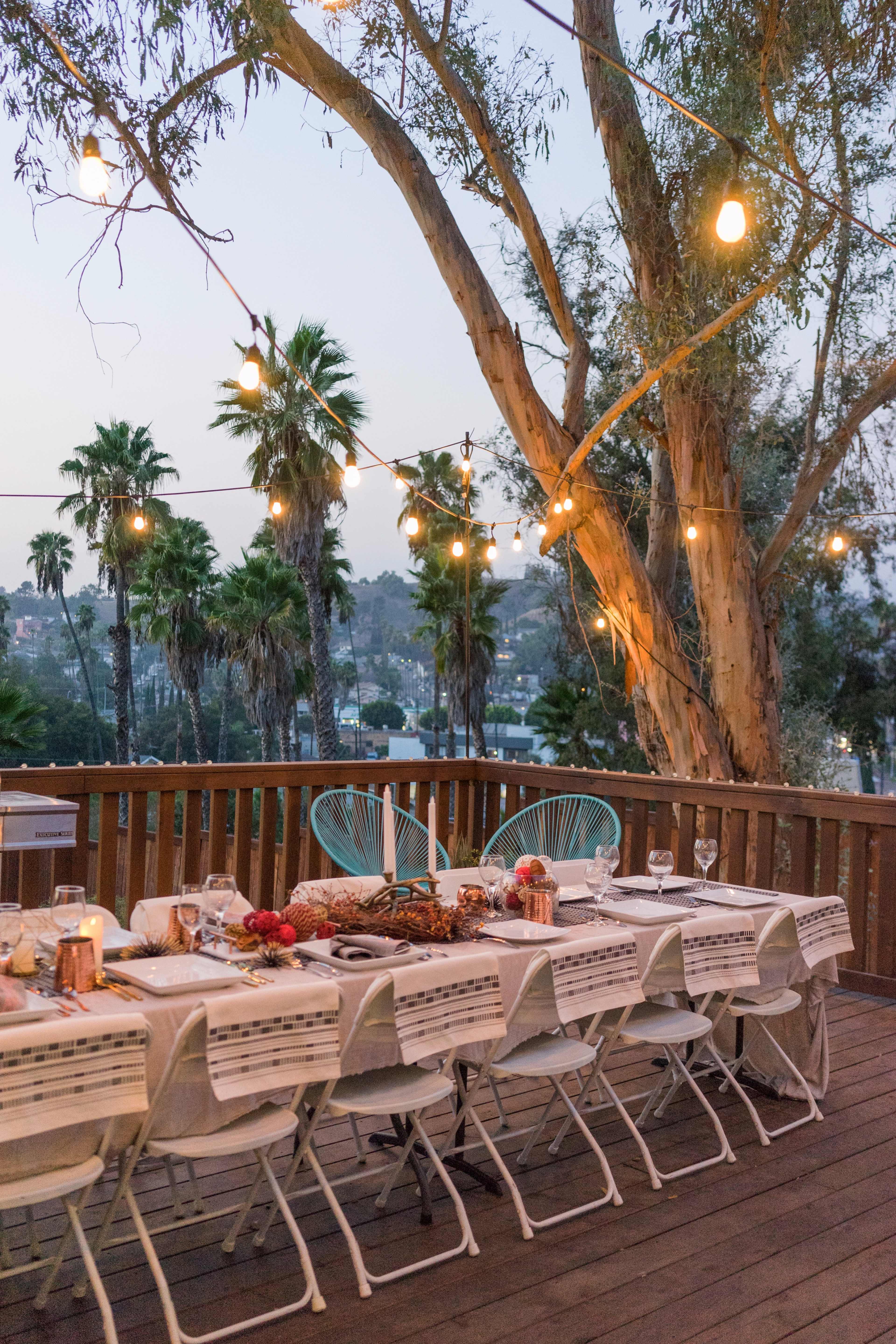 A long dining table is set on a deck under string lights, surrounded by palm trees and overlooking a scenic view at dusk.