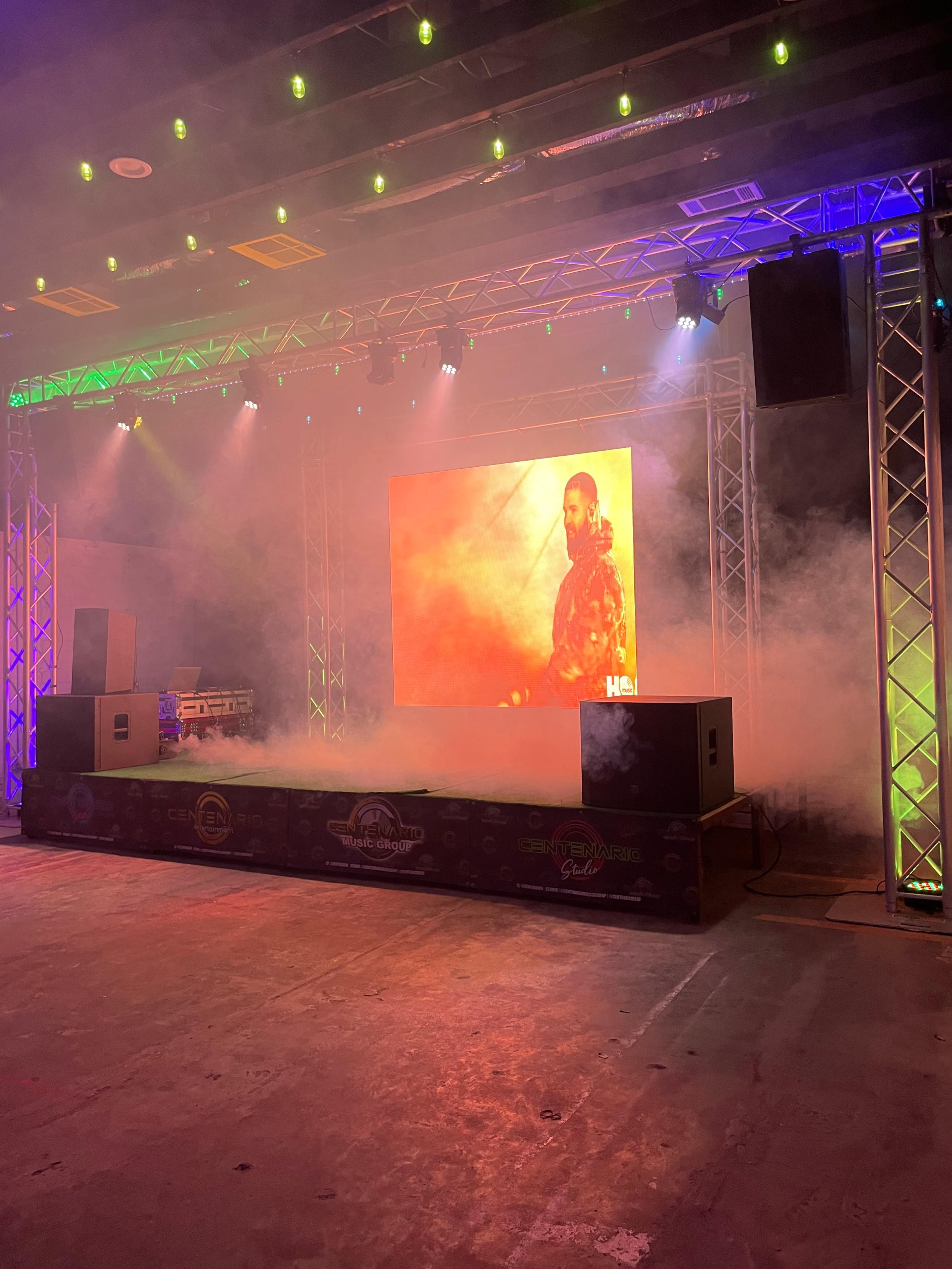 A stage setup features illuminated trussing, smoke effects, and a large backdrop displaying a performer.
