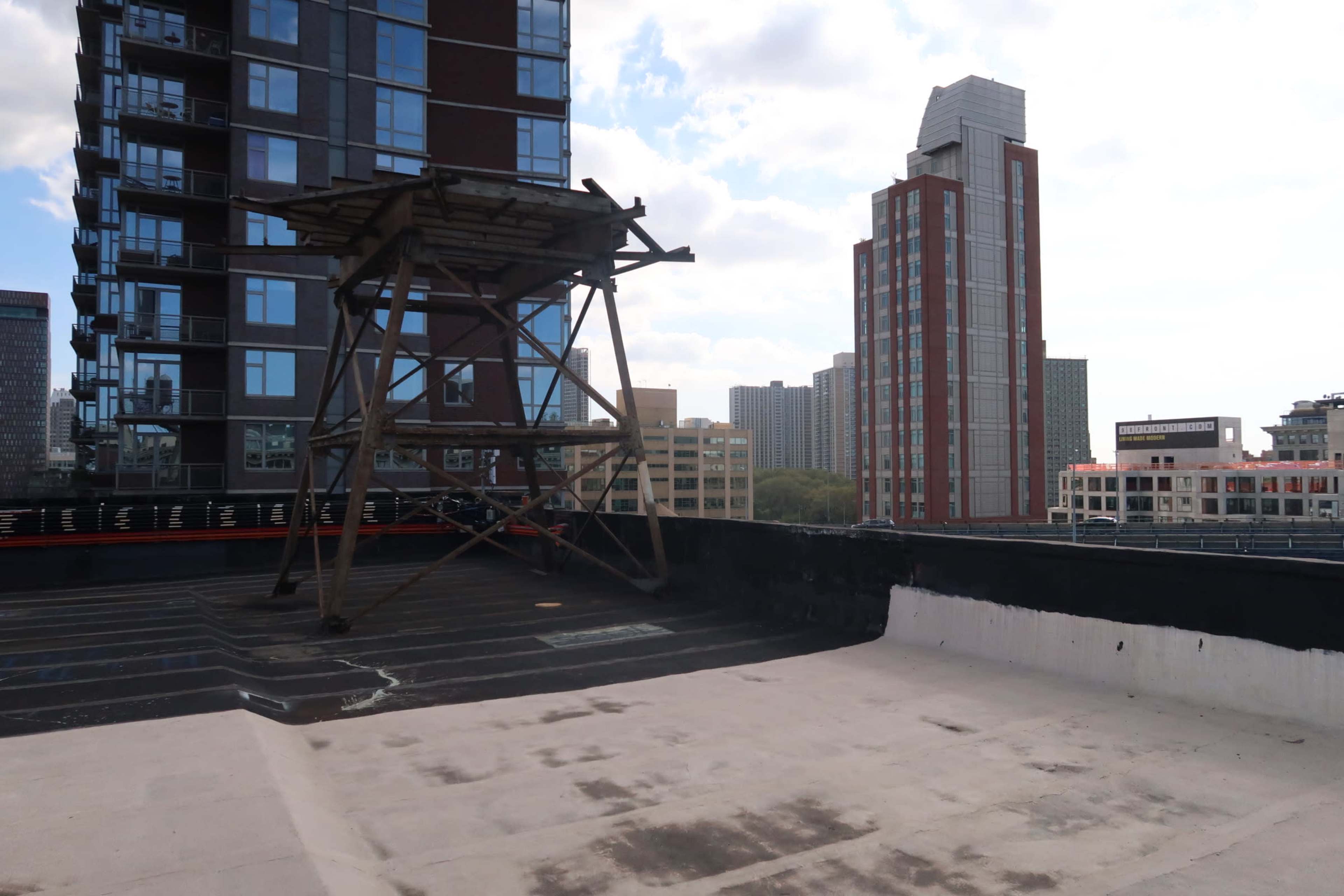 A concrete rooftop features a wooden frame structure and overlooks nearby high-rise buildings against a cloudy sky.