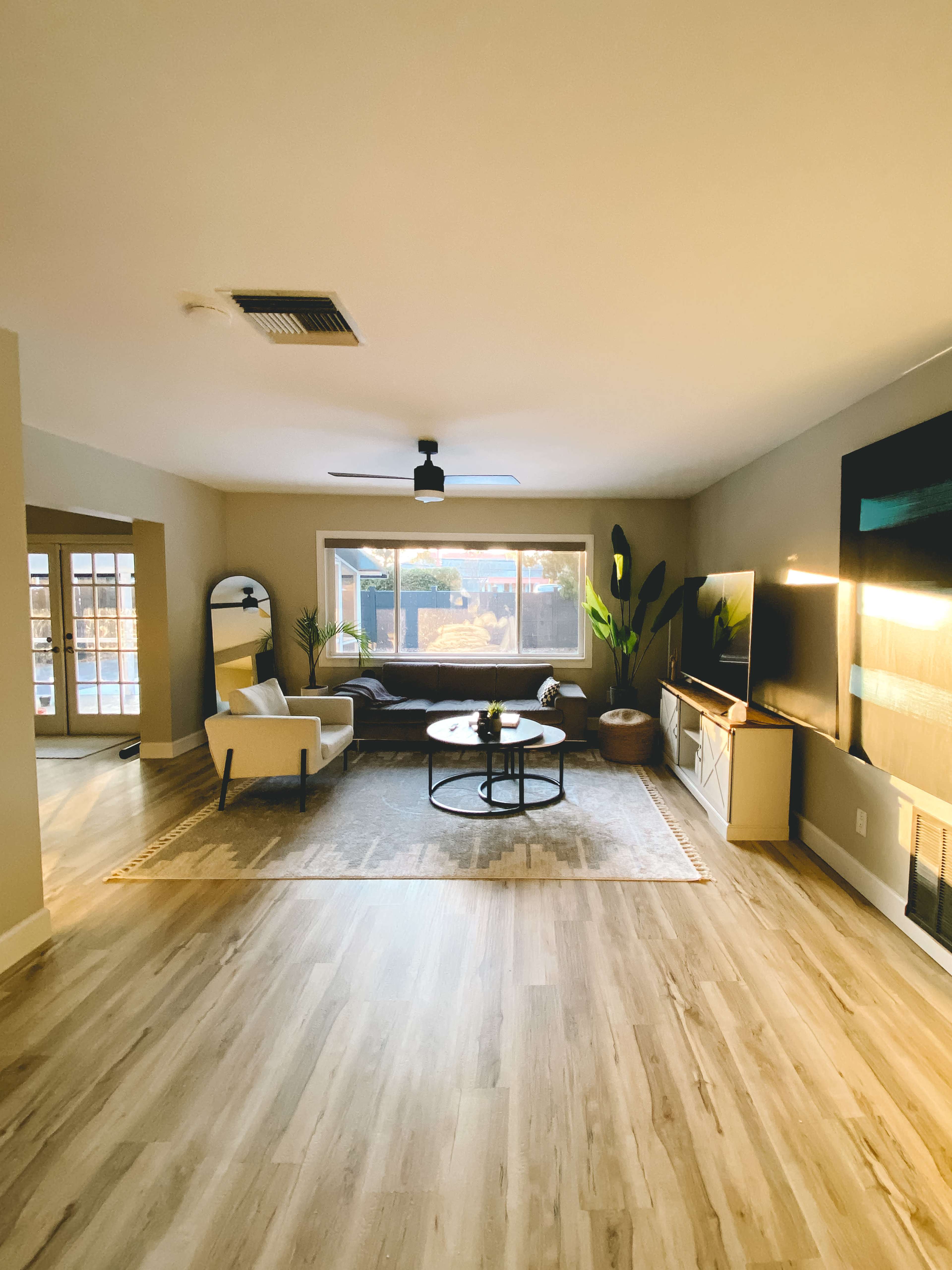 A spacious living room with a seating area, a round coffee table, a large window, and a television on a cabinet against a wall.