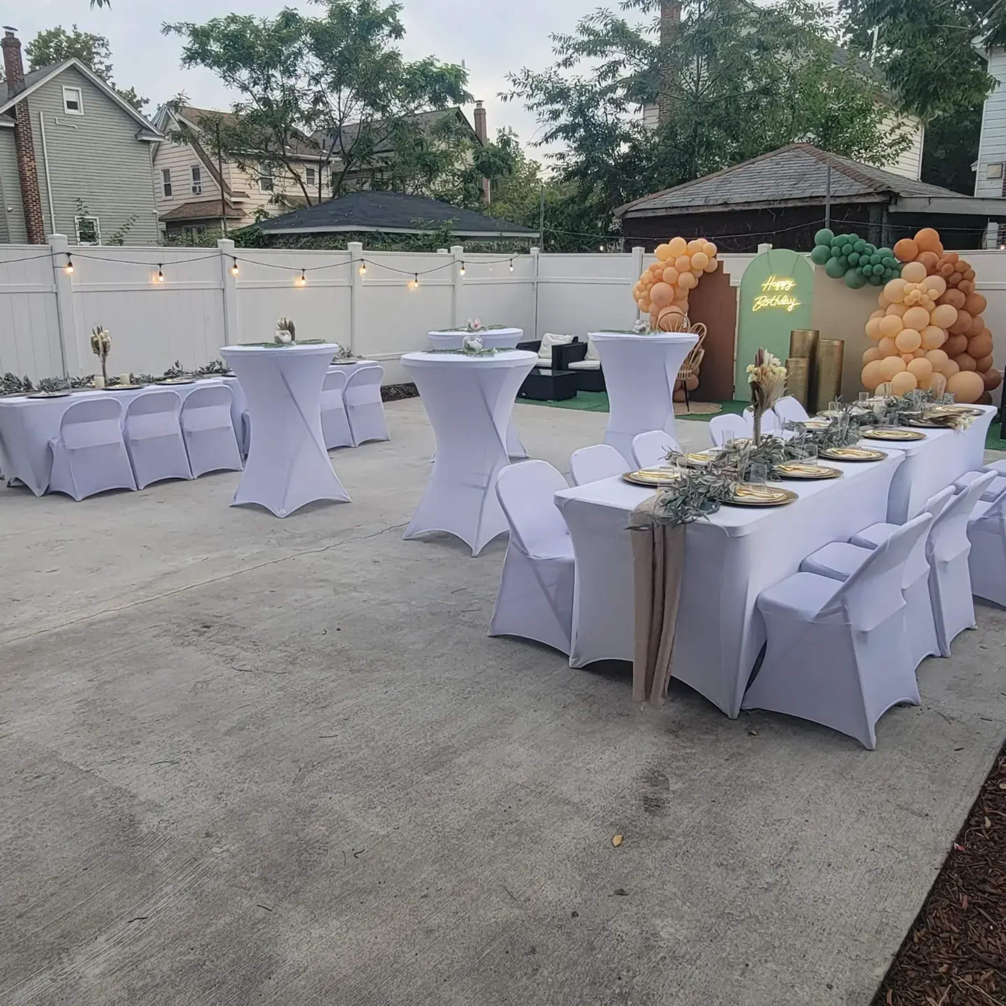 A backyard event setup features tables covered in white linens, decorated with gold accents and greenery, surrounded by a colorful balloon arch and string lights.