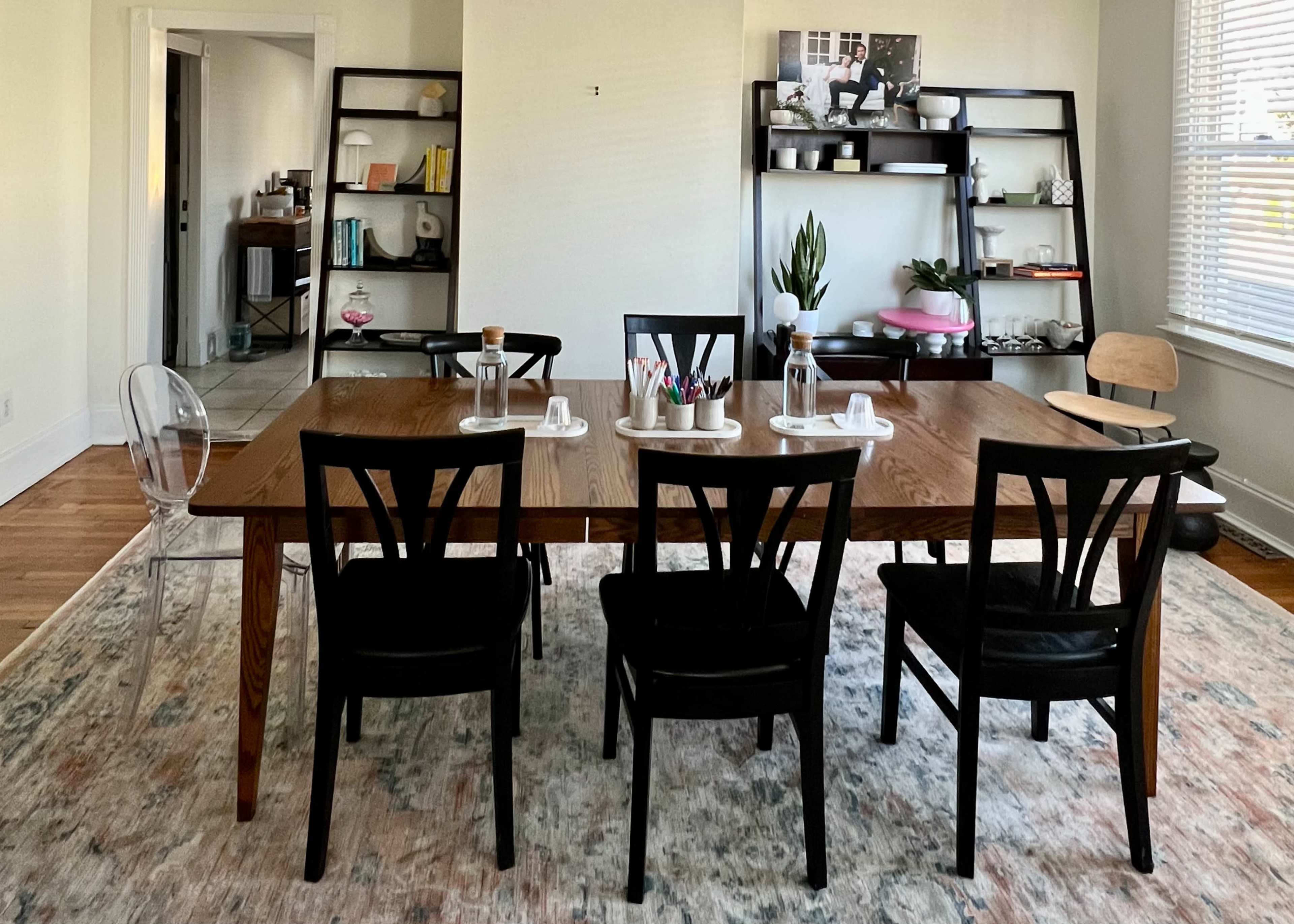 A wooden dining table with six black chairs and decorative items is arranged in a bright, minimalist room with shelves displaying plants and books.