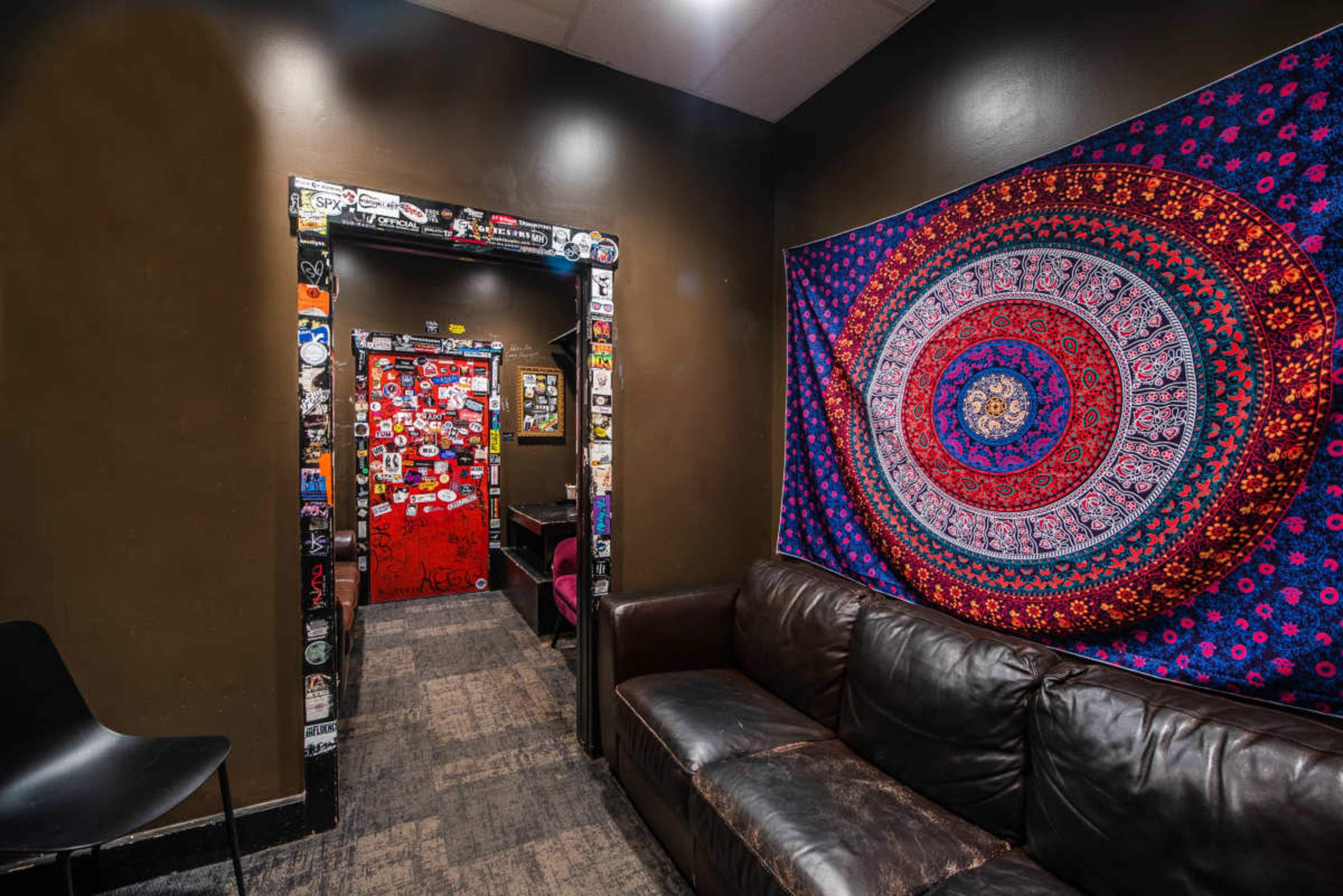 The image shows a dimly lit room with brown walls, a dark couch, and a colorful tapestry hanging next to a door covered in various stickers.
