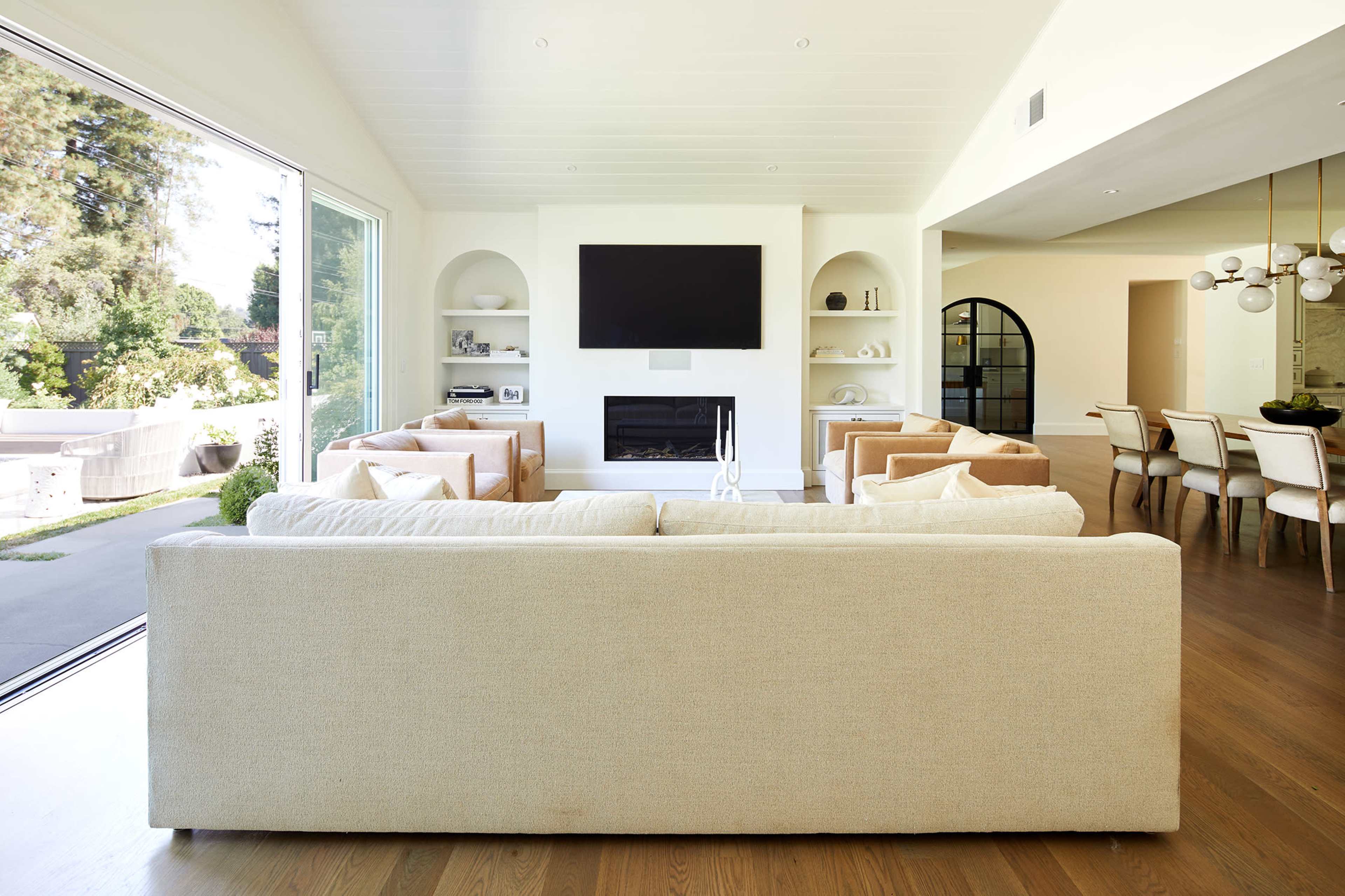 A modern living room with a large sofa facing a wall-mounted television, surrounded by two armchairs and an open dining area in the background.