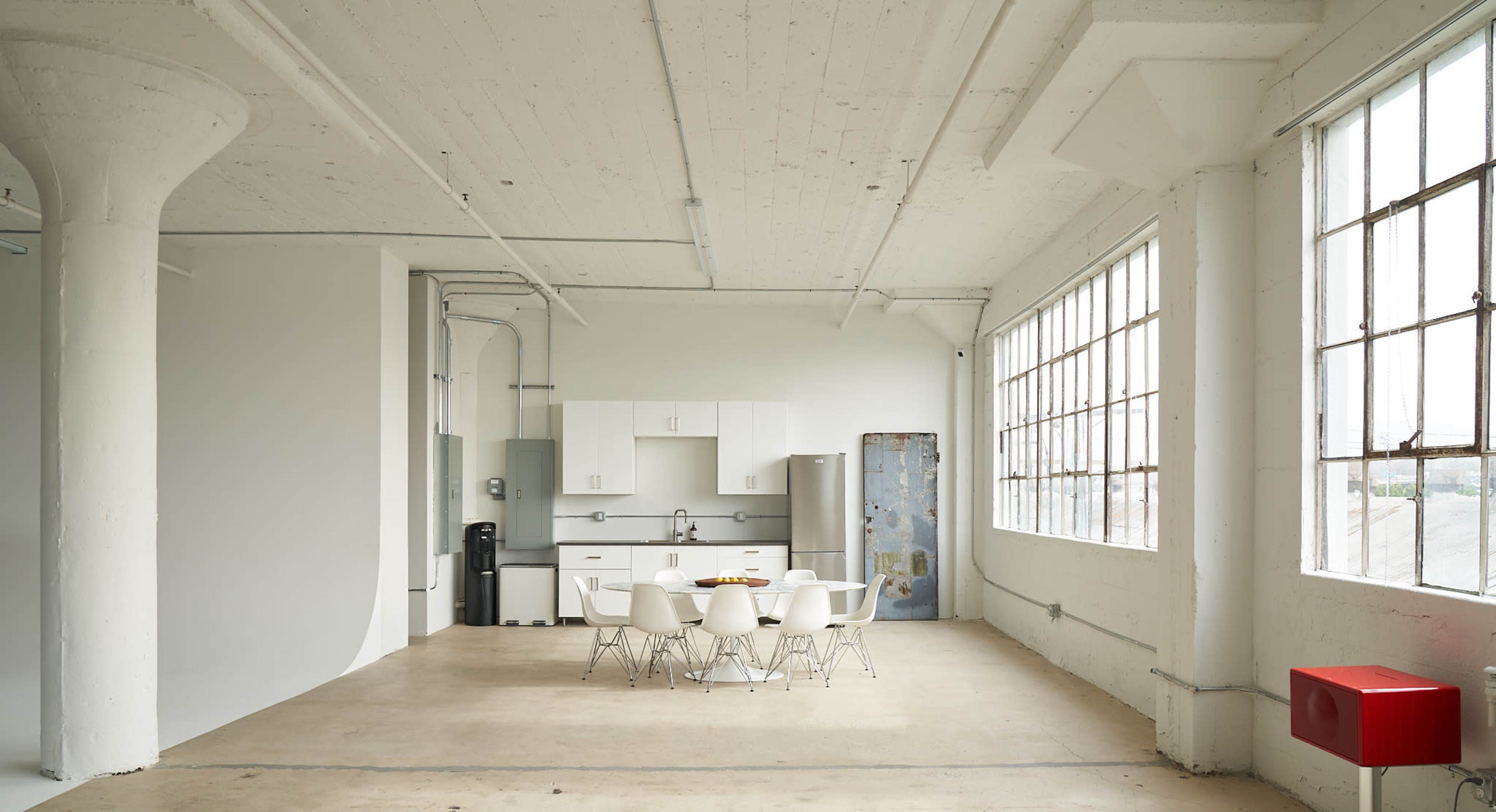 The image shows a modern, minimalist kitchen and dining area with a round table surrounded by white chairs, large windows allowing natural light, and basic kitchen cabinetry.