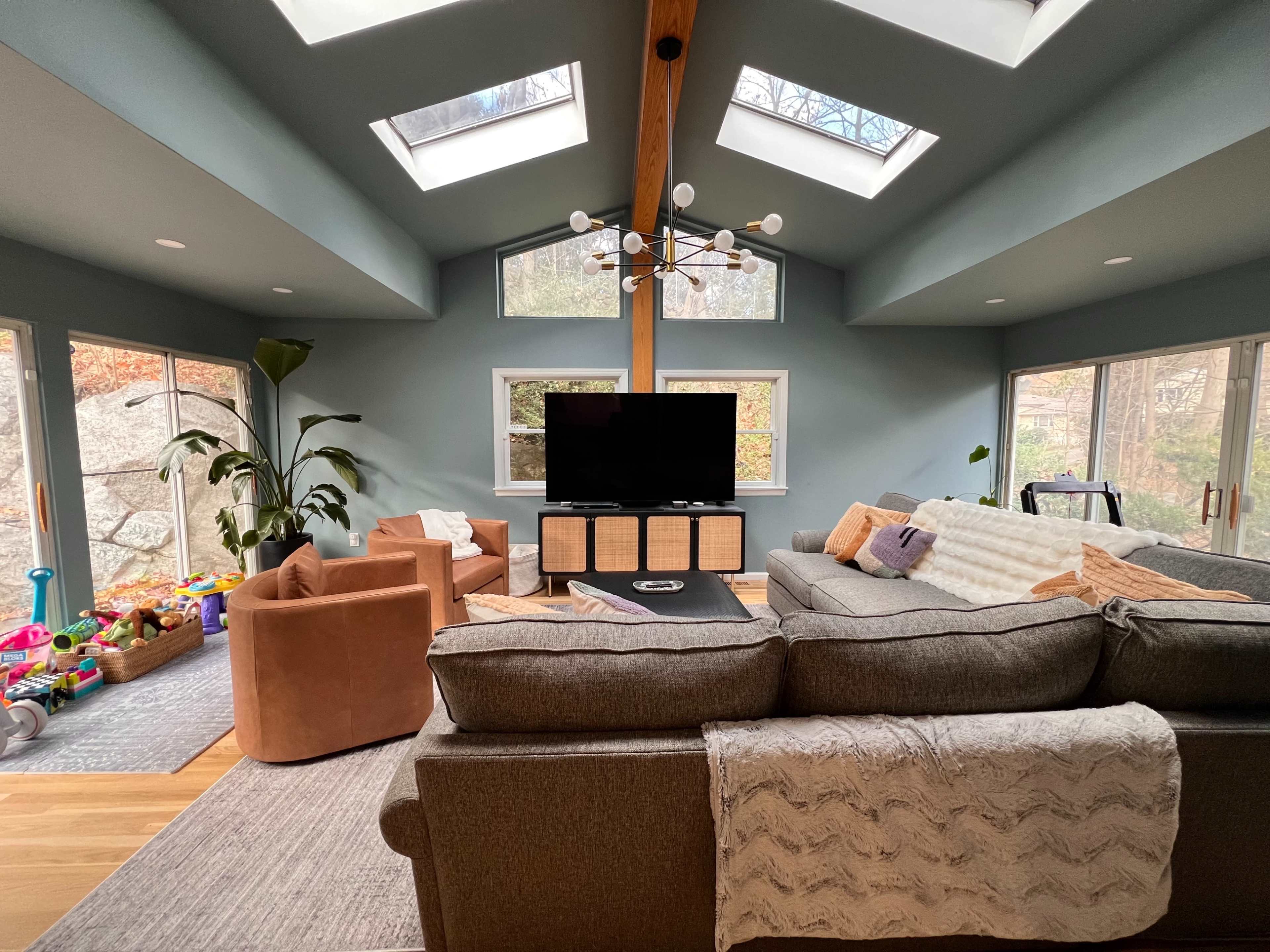 A spacious living room features a sectional sofa, armchairs, a television on a wooden cabinet, and large windows that allow natural light.