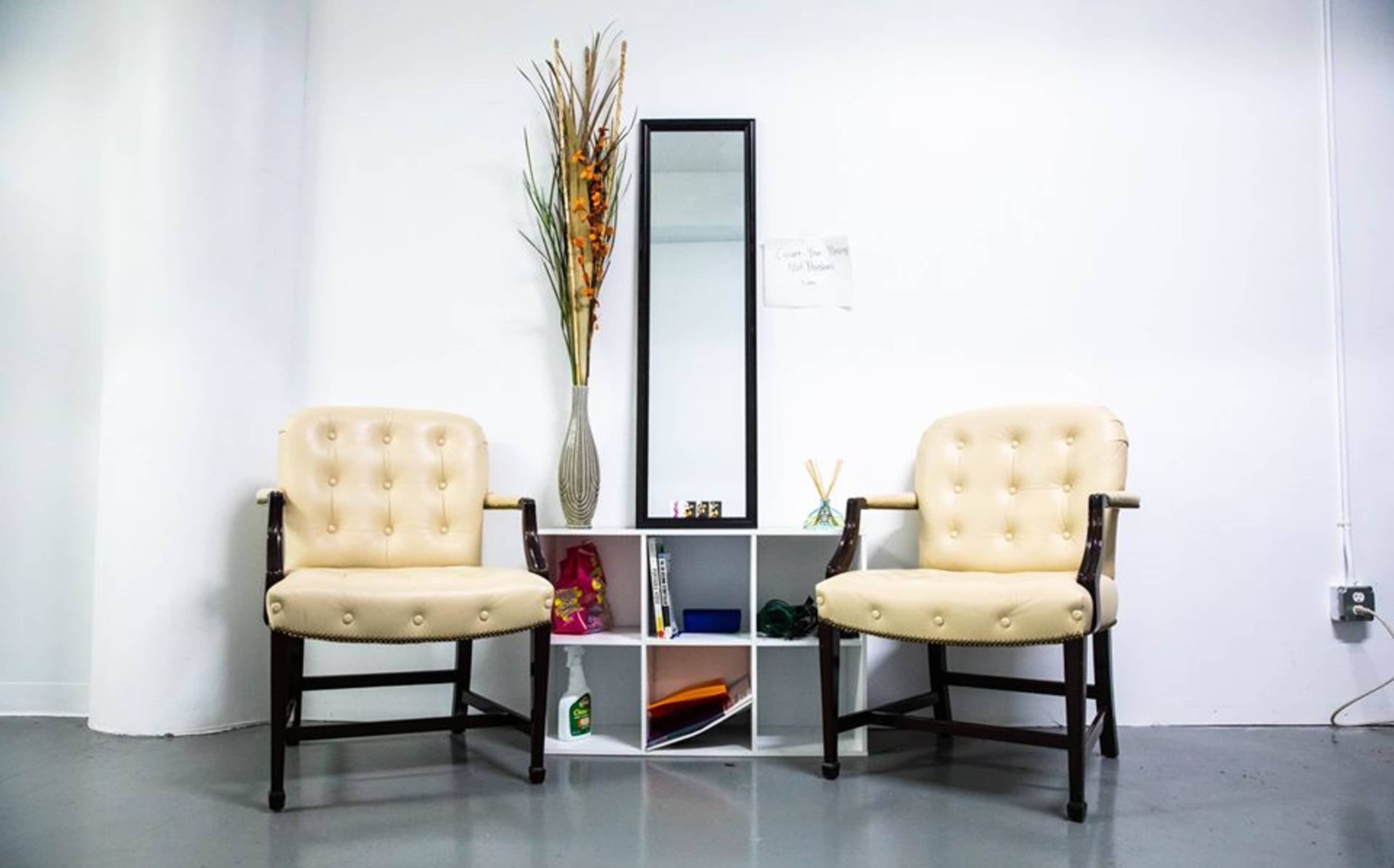 Two beige upholstered chairs facing a small shelf with a mirror and decorative items in a bright, minimally decorated room.