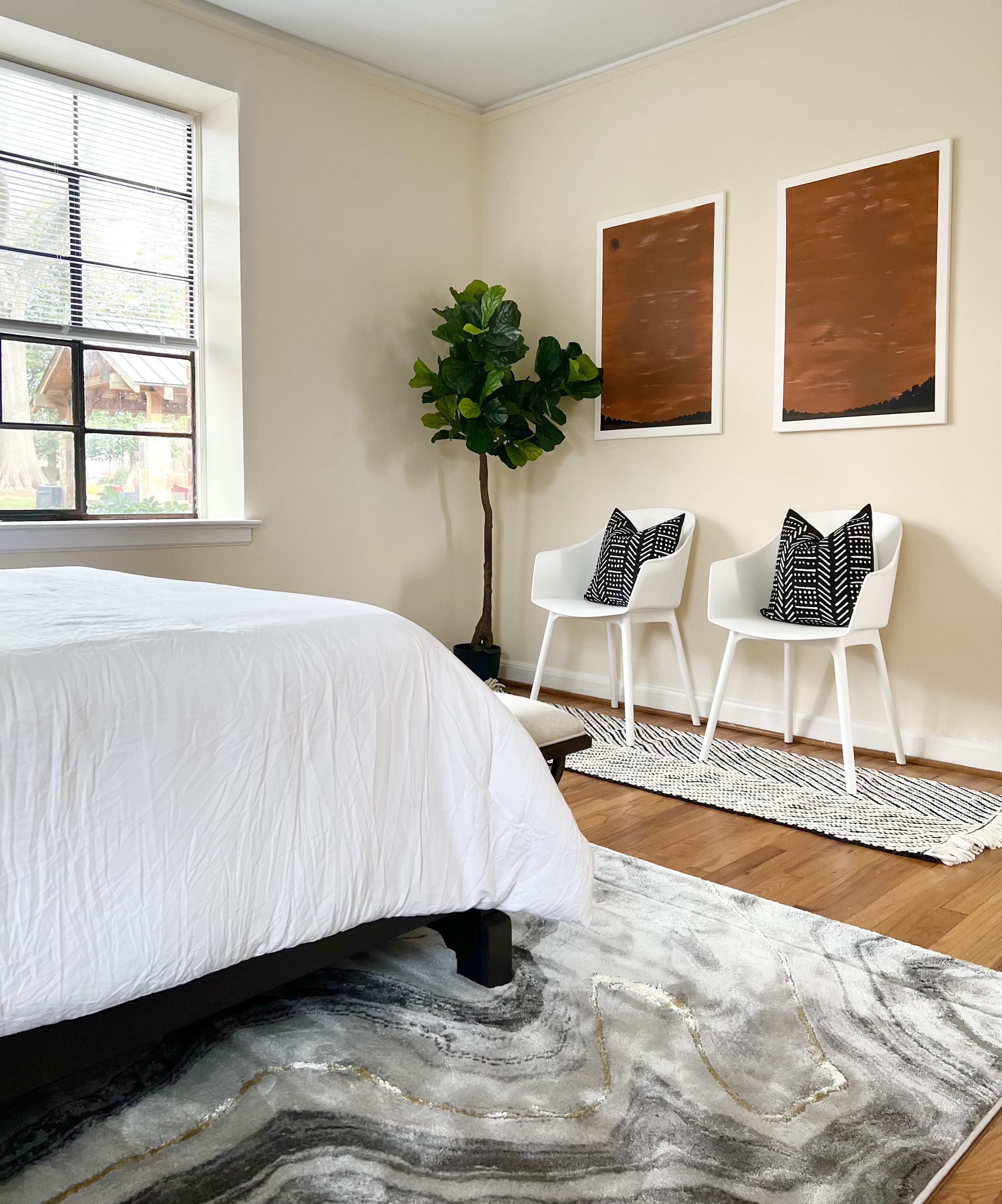The room features a white bedspread, two white chairs with patterned cushions, a tall plant, and two framed artworks hanging on the wall.
