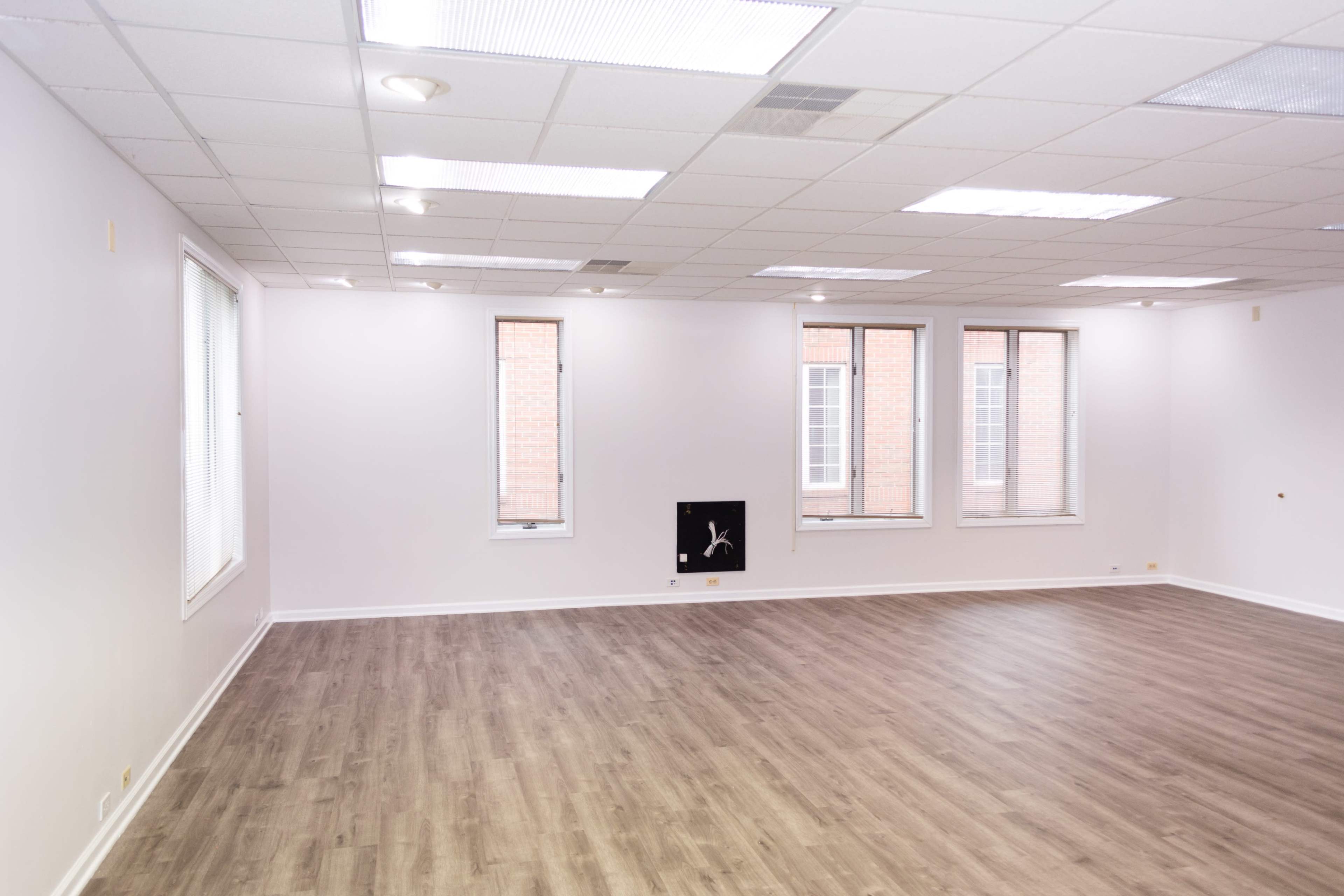 An empty room with wooden flooring and three windows, featuring plain white walls and a small wall-mounted object.