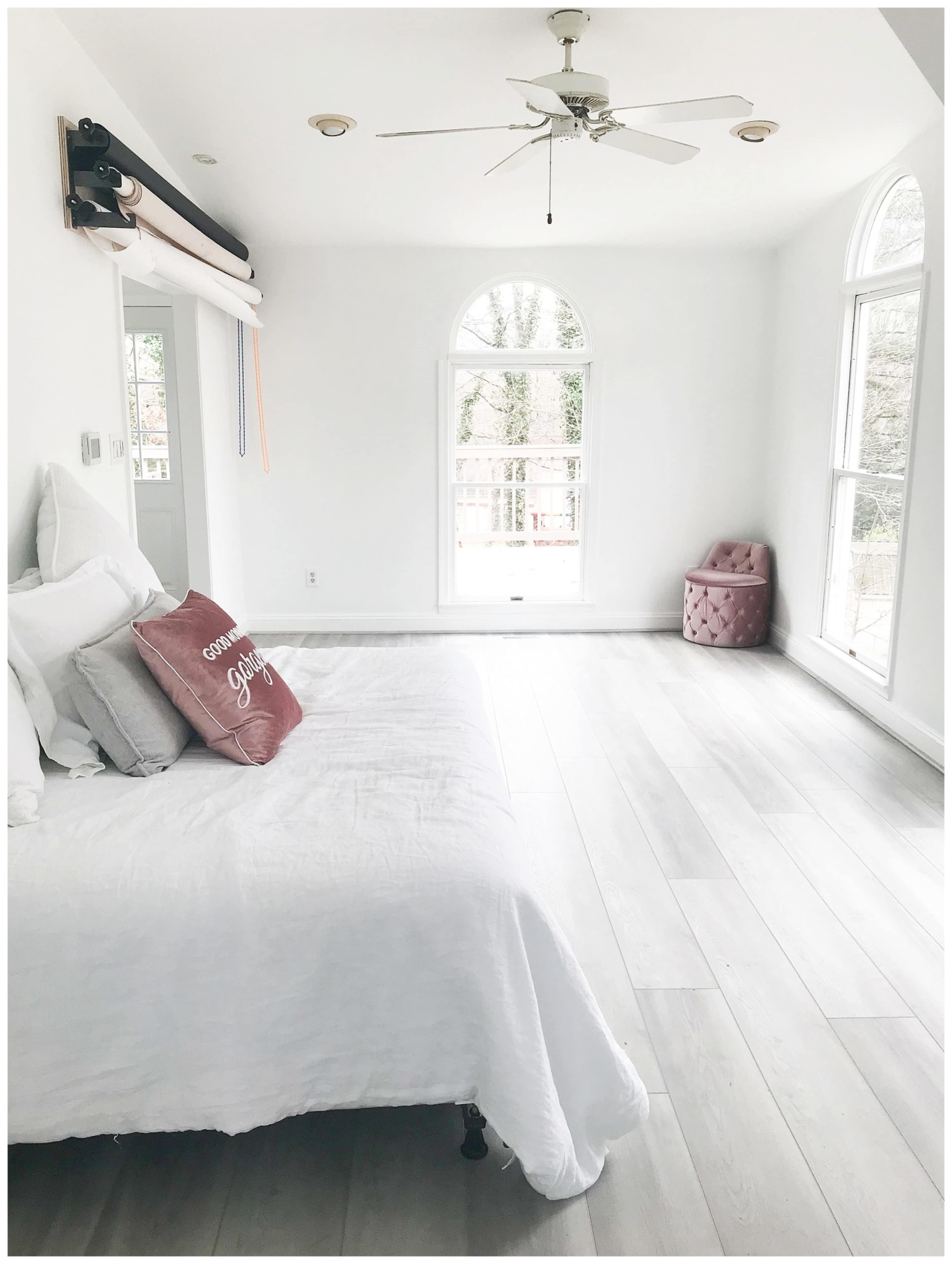 A well-lit room features a white bed with a decorative pillow, a pink upholstered chair, and large windows overlooking a balcony.
