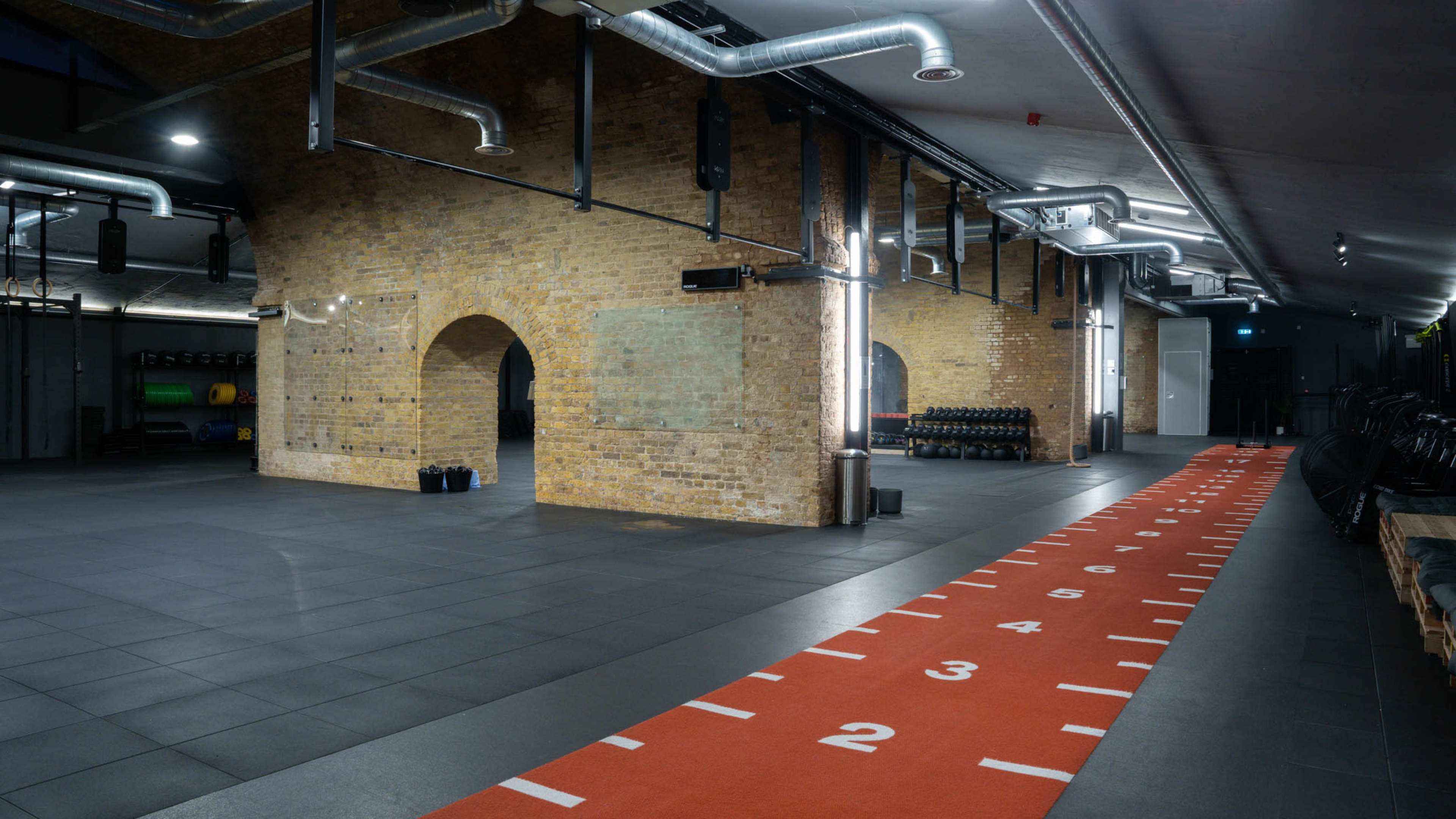A spacious gym interior featuring exposed brick walls, a gray tiled floor, and an orange track marked with numbers on one side.