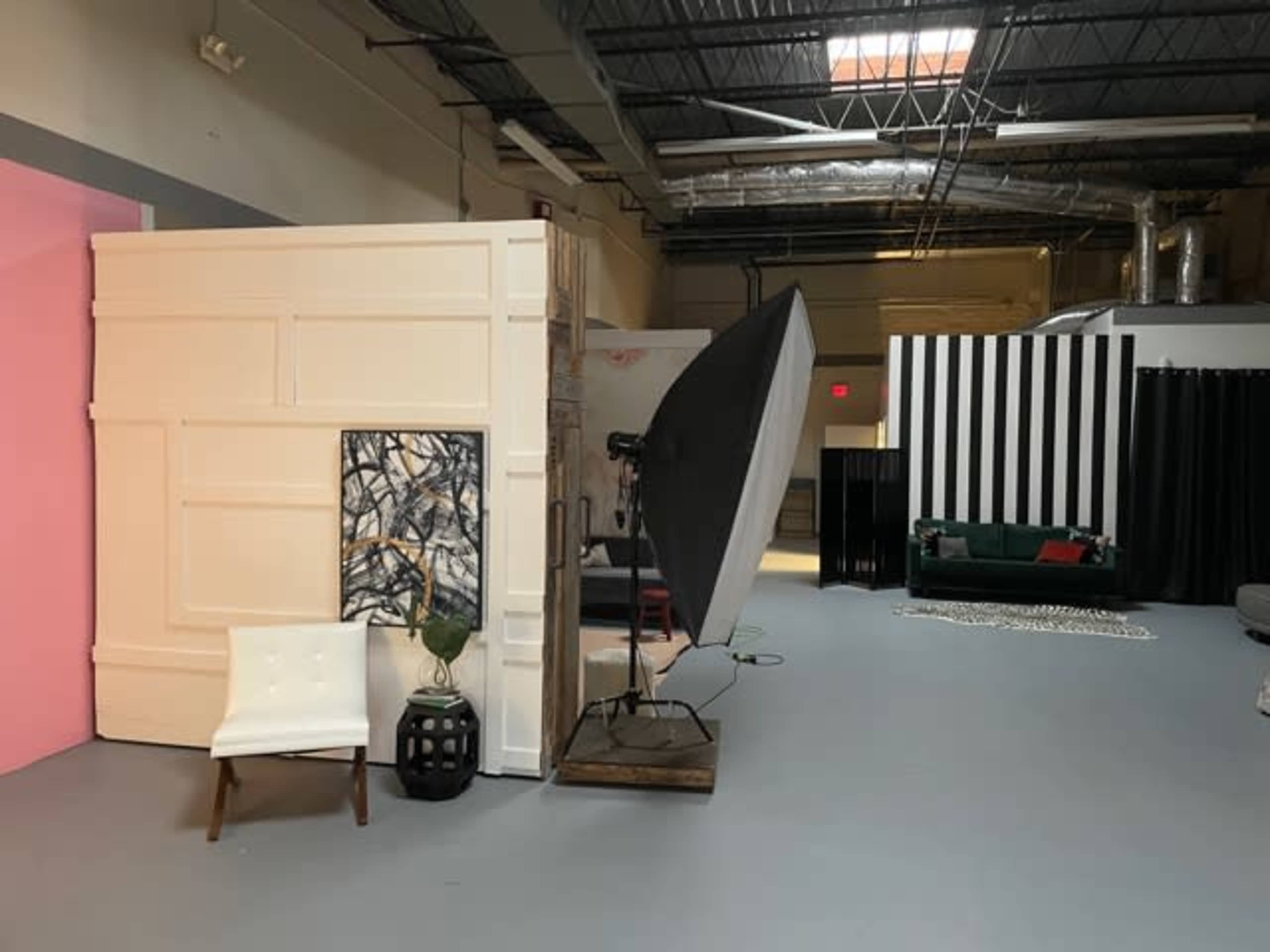 A spacious studio interior with various backdrops, a large softbox light, and a mix of furniture arrangements, including a chair and a sofa.