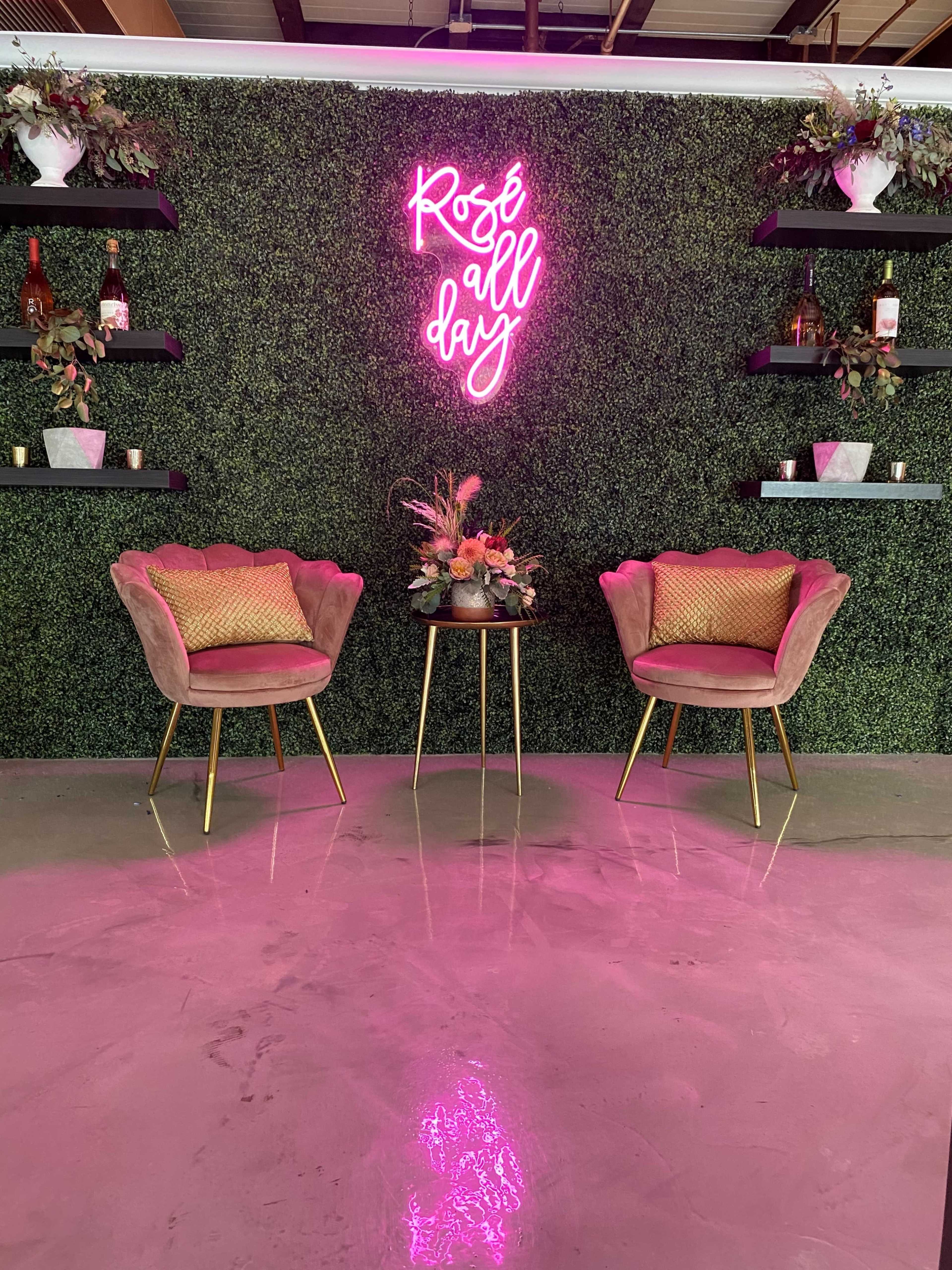 The scene features two pink chairs with gold legs set against a green foliage wall, adorned with shelves holding decorative items and a neon sign that reads "Rosé all day."