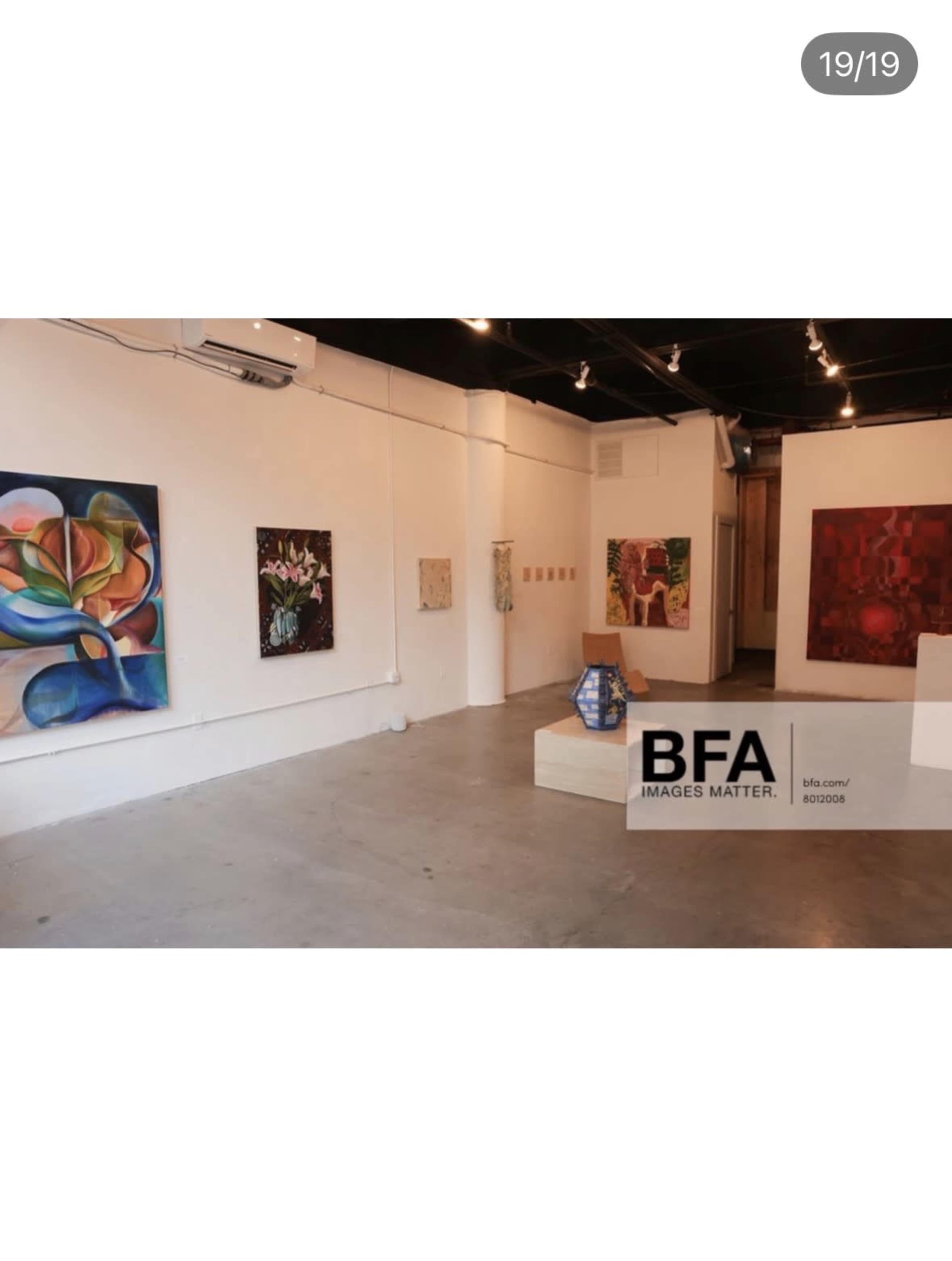 The image shows an art gallery with several colorful paintings displayed on the walls and a small sculpture on a pedestal in the center.