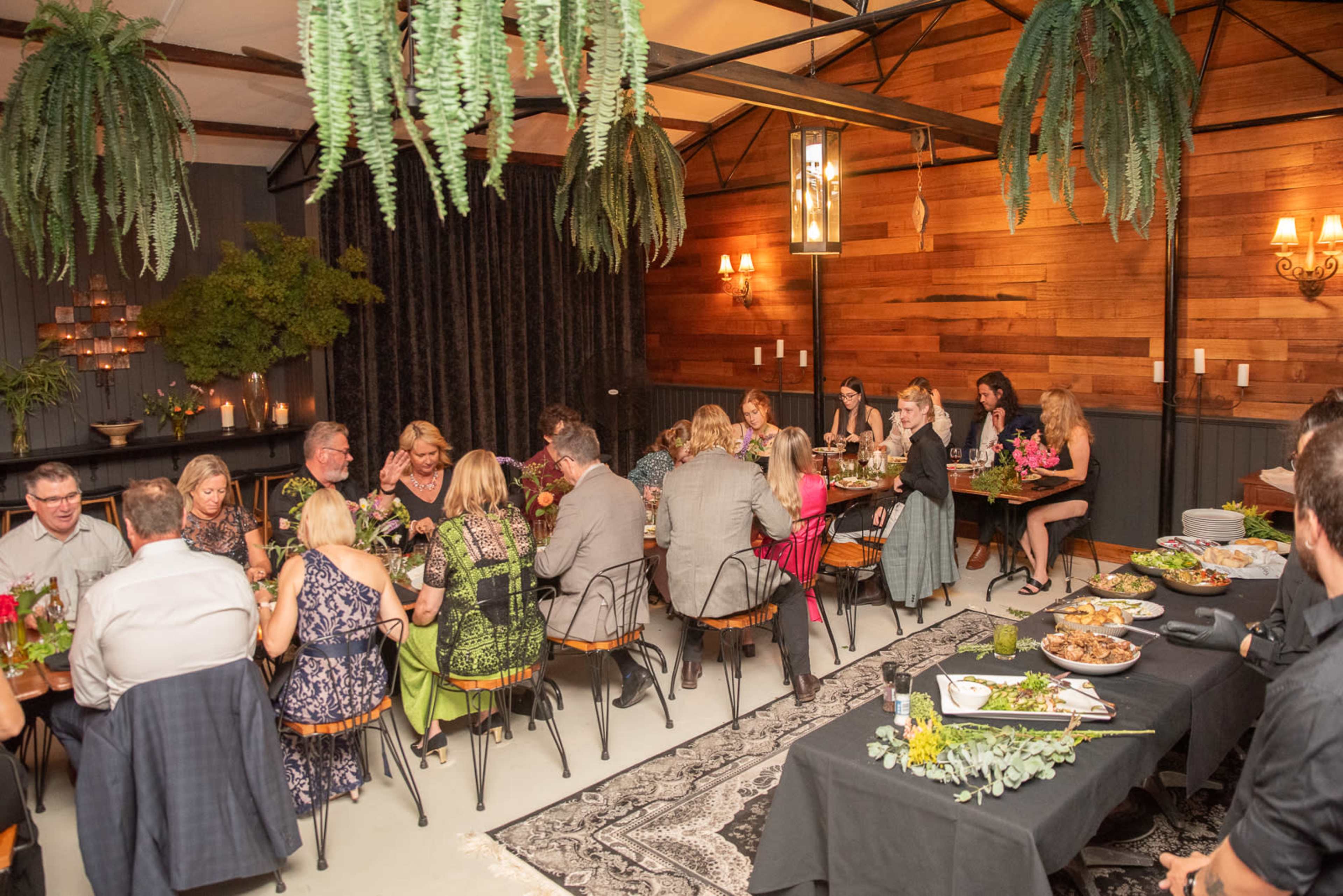 A group of people dine together in a warmly lit, rustic room adorned with wooden walls and greenery.