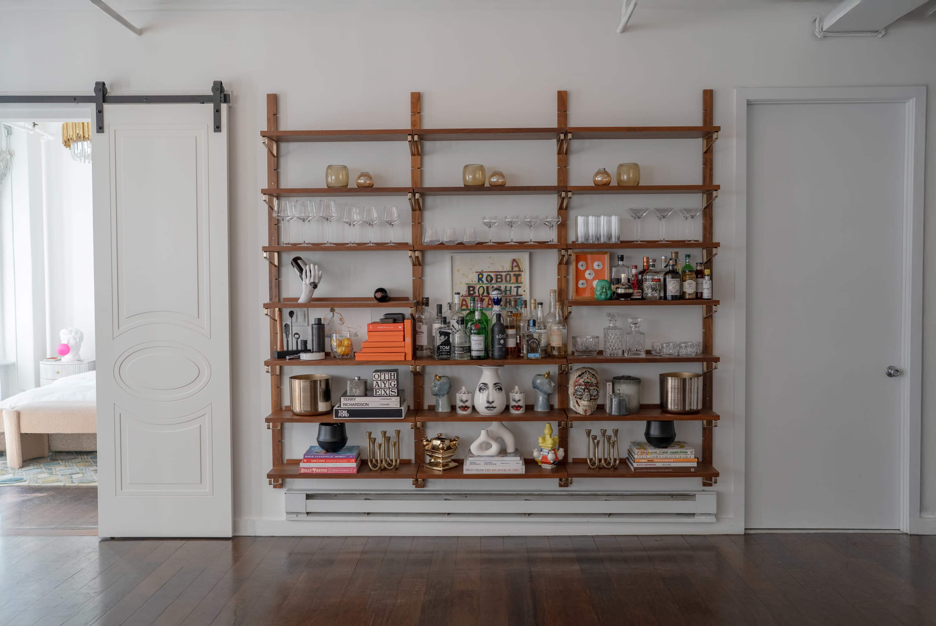 The image shows a wooden wall shelf unit filled with a variety of glassware, bottles, decorative items, and books, set against a white wall in a well-lit room.