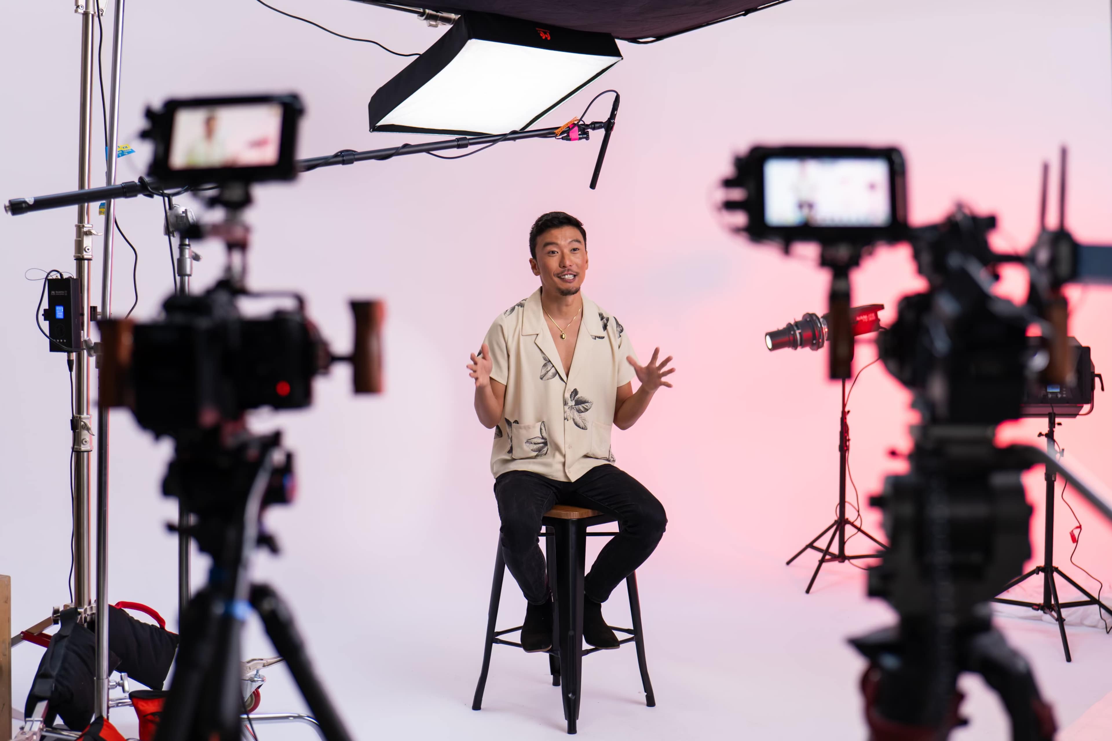 A person is seated on a stool in a studio, surrounded by several cameras on tripods, with bright light sources overhead.