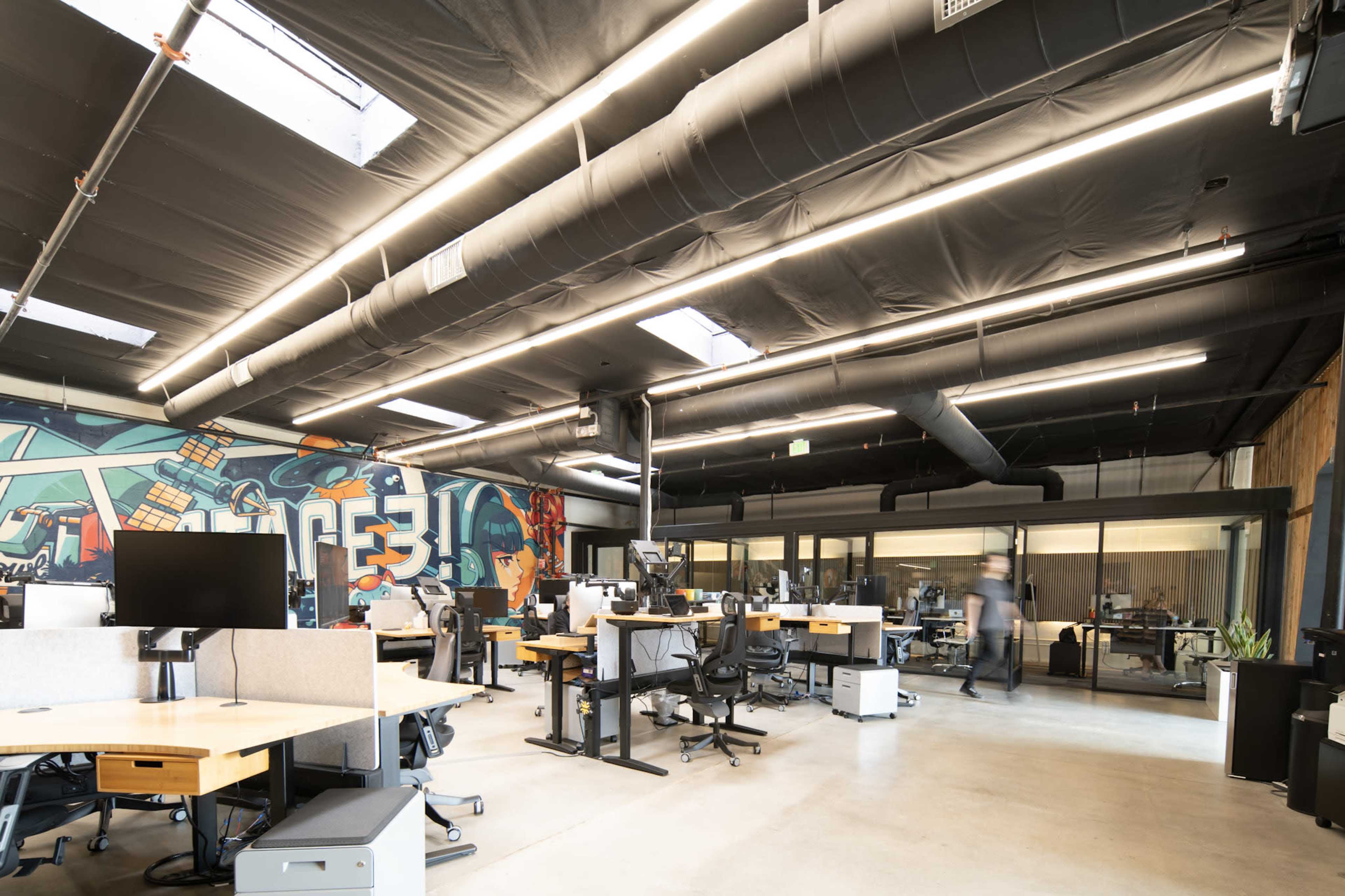 The image shows a modern office space featuring multiple workstations, a colorful mural on one wall, and large windows allowing light into the area.