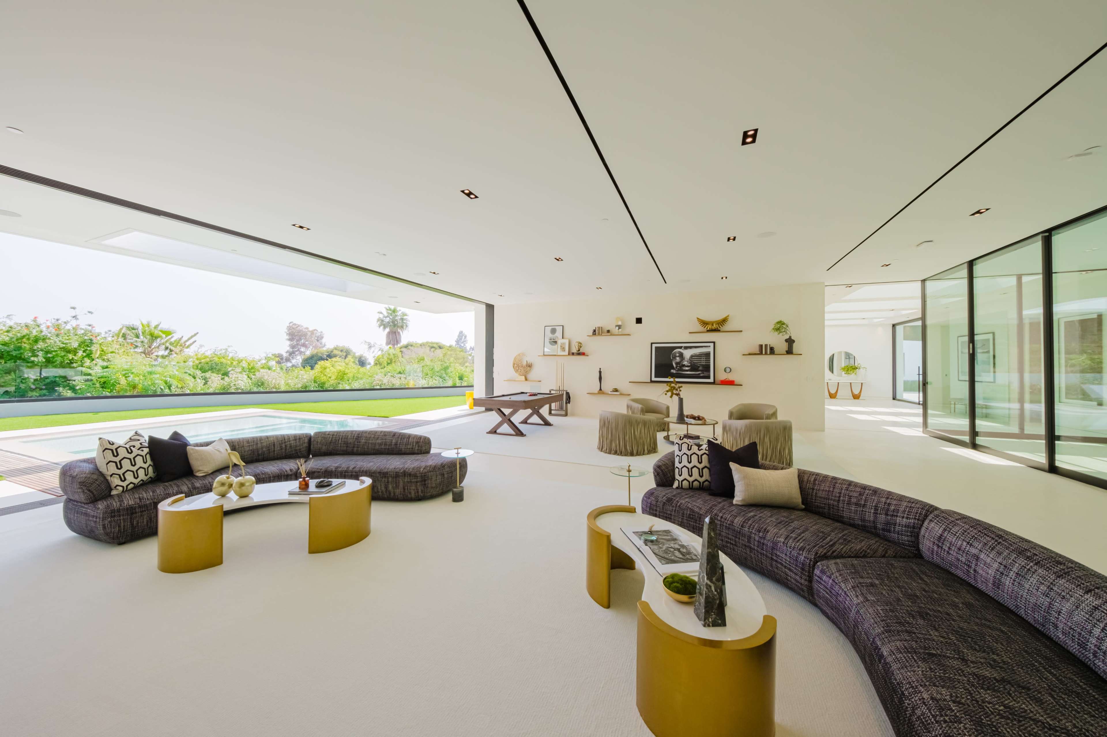 A modern living room with large windows, featuring curved sectional sofas and minimalistic decor, overlooking a landscaped outdoor area.