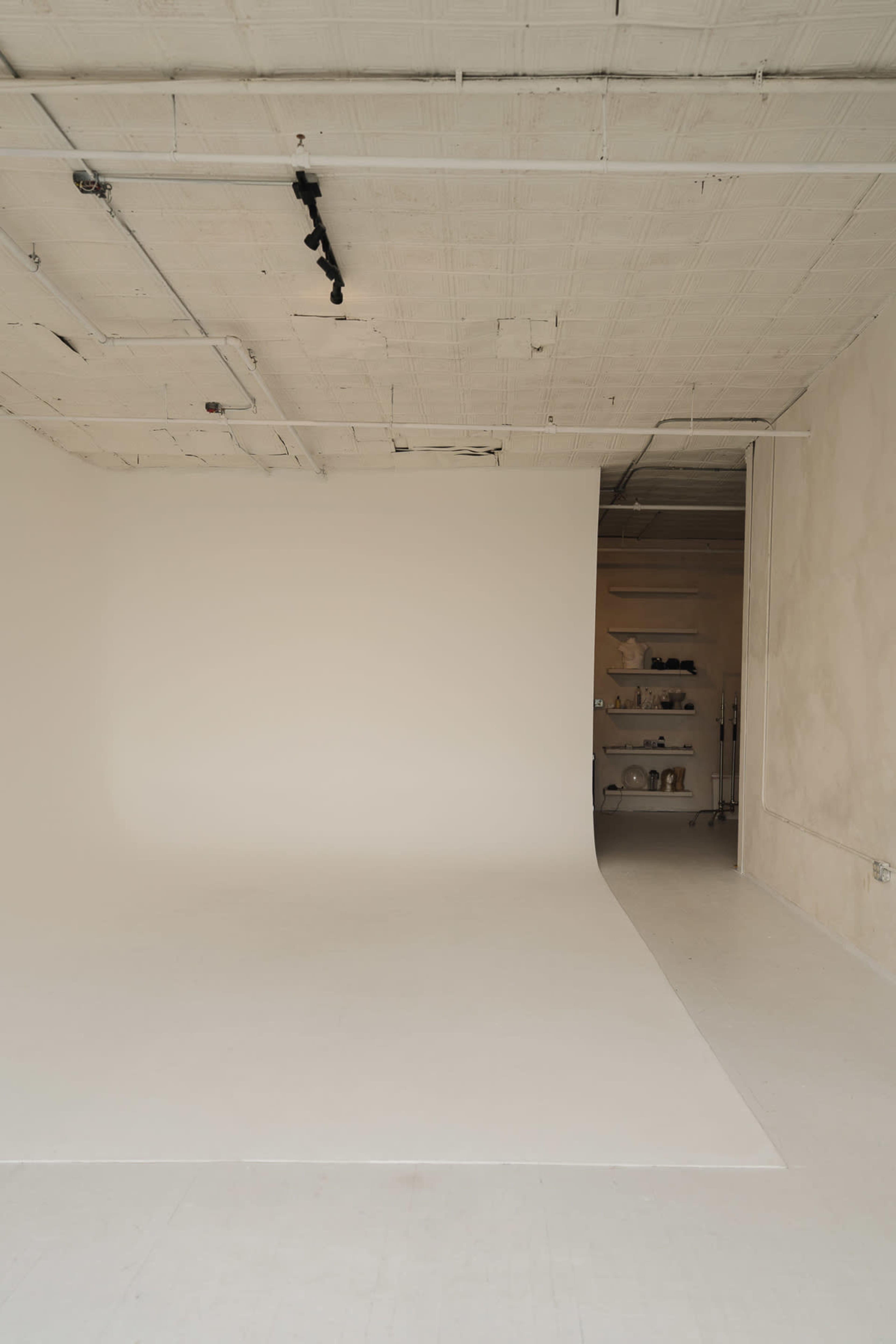 The image shows a spacious, minimalist photography studio featuring a blank white backdrop and a partially open room with shelves in the background.