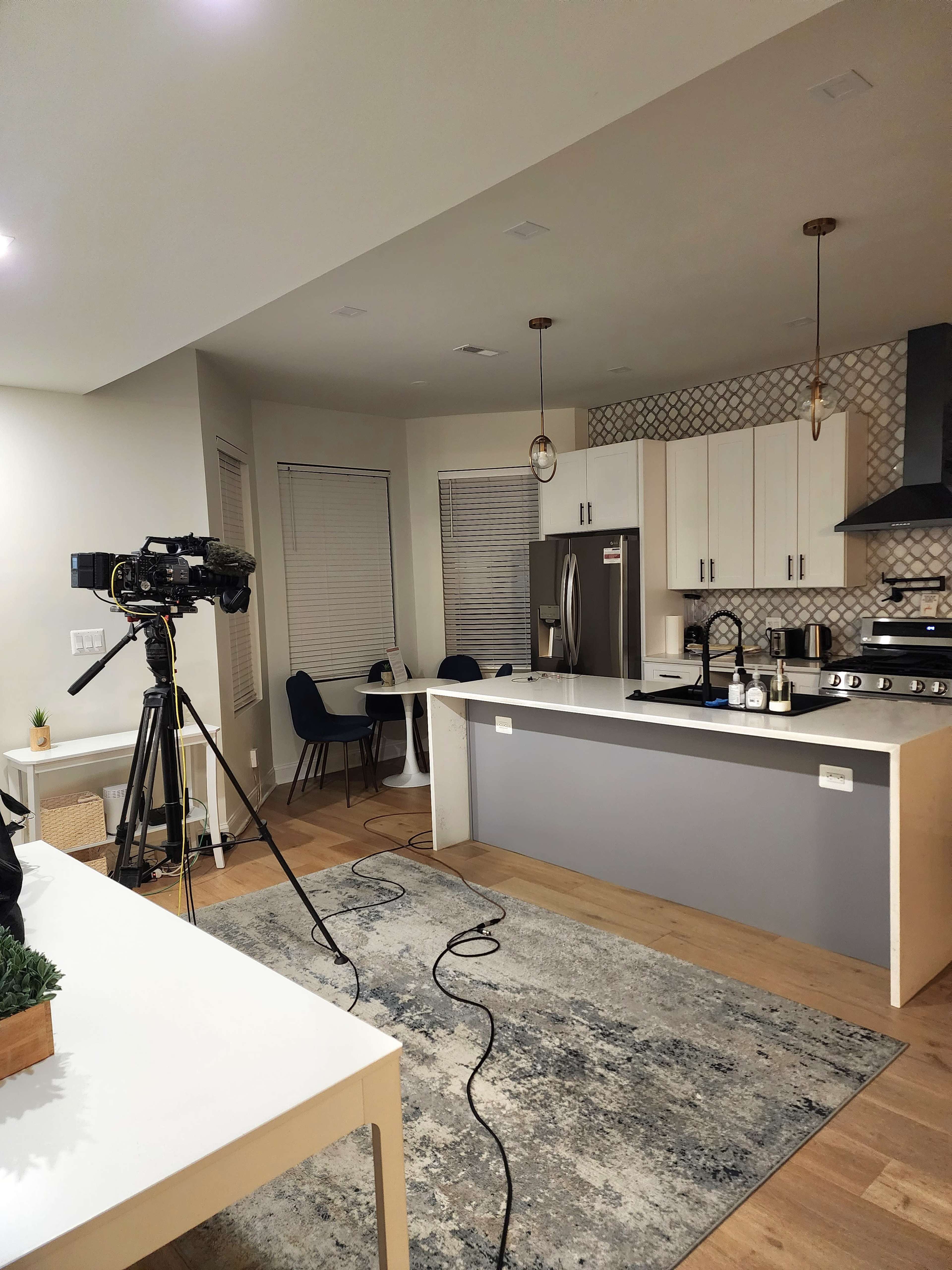 A kitchen is set up for filming, featuring a camera on a tripod, a dining table with chairs, and modern appliances.