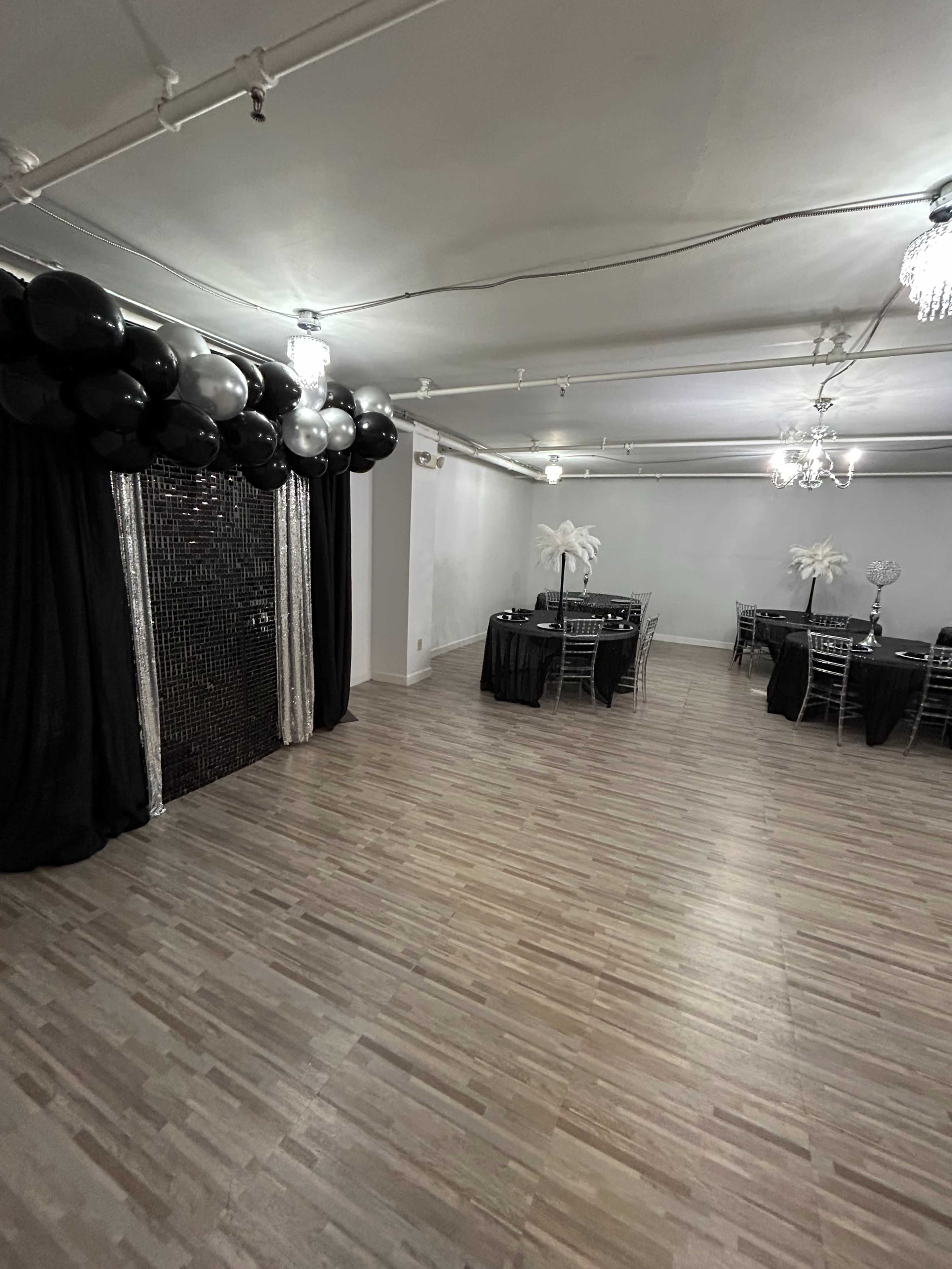 A spacious event room decorated with black and silver balloons, black tablecloths, and feather centerpieces.