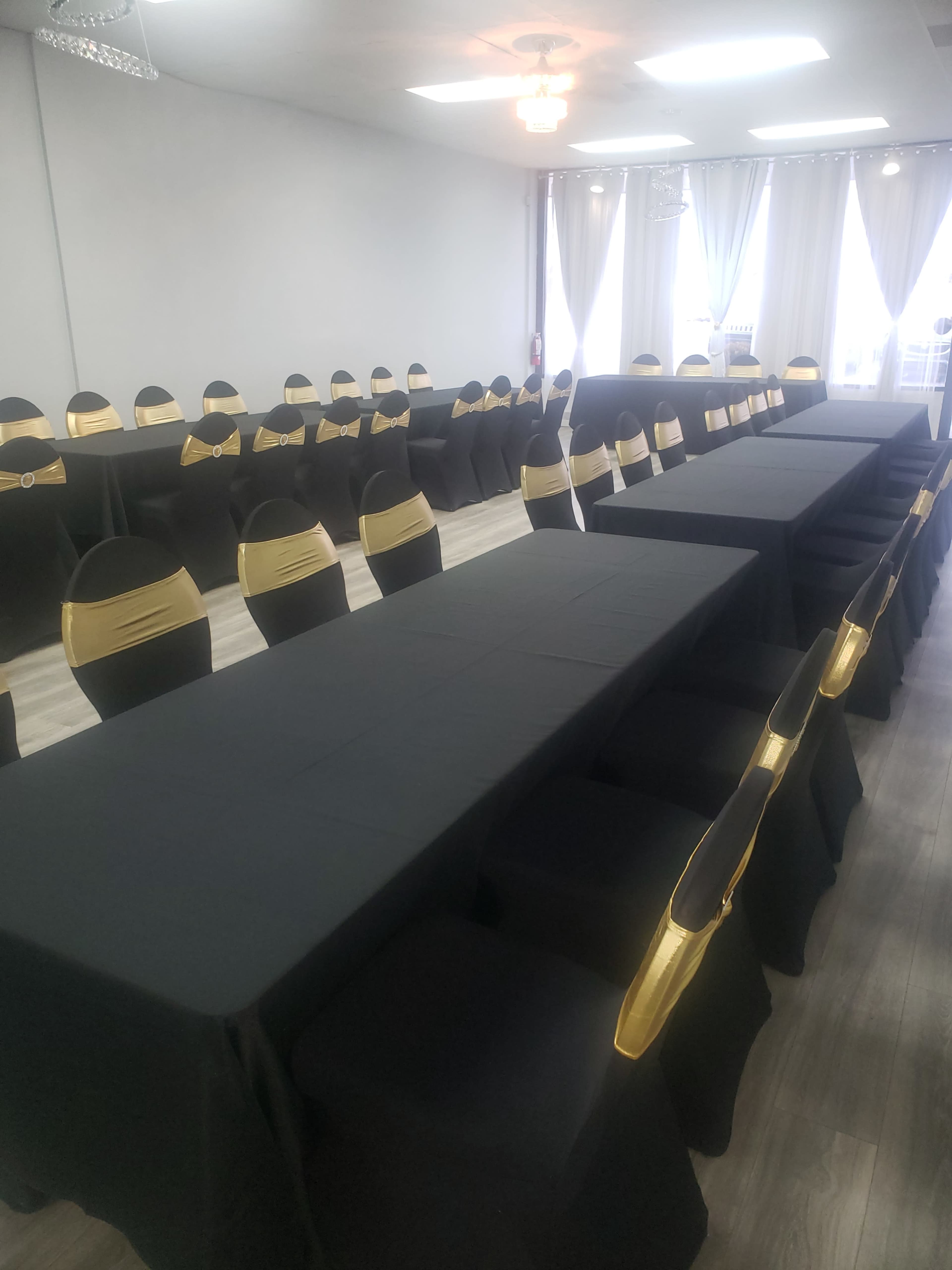 A spacious banquet room features long tables covered with black tablecloths and chairs adorned with gold sashes.