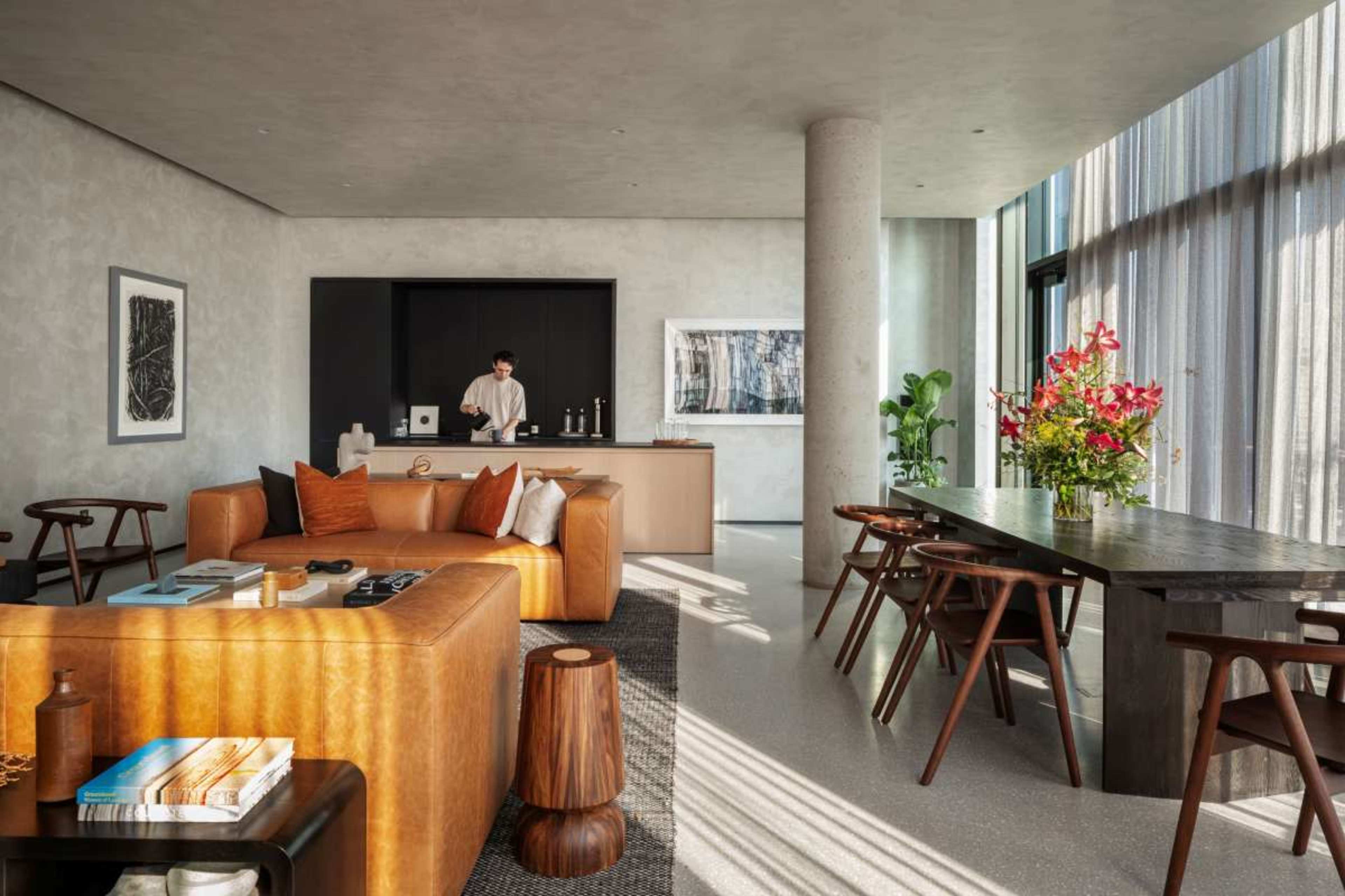 The image shows a modern living space with a leather sofa, wooden dining table, and a person preparing drinks in the kitchen area.
