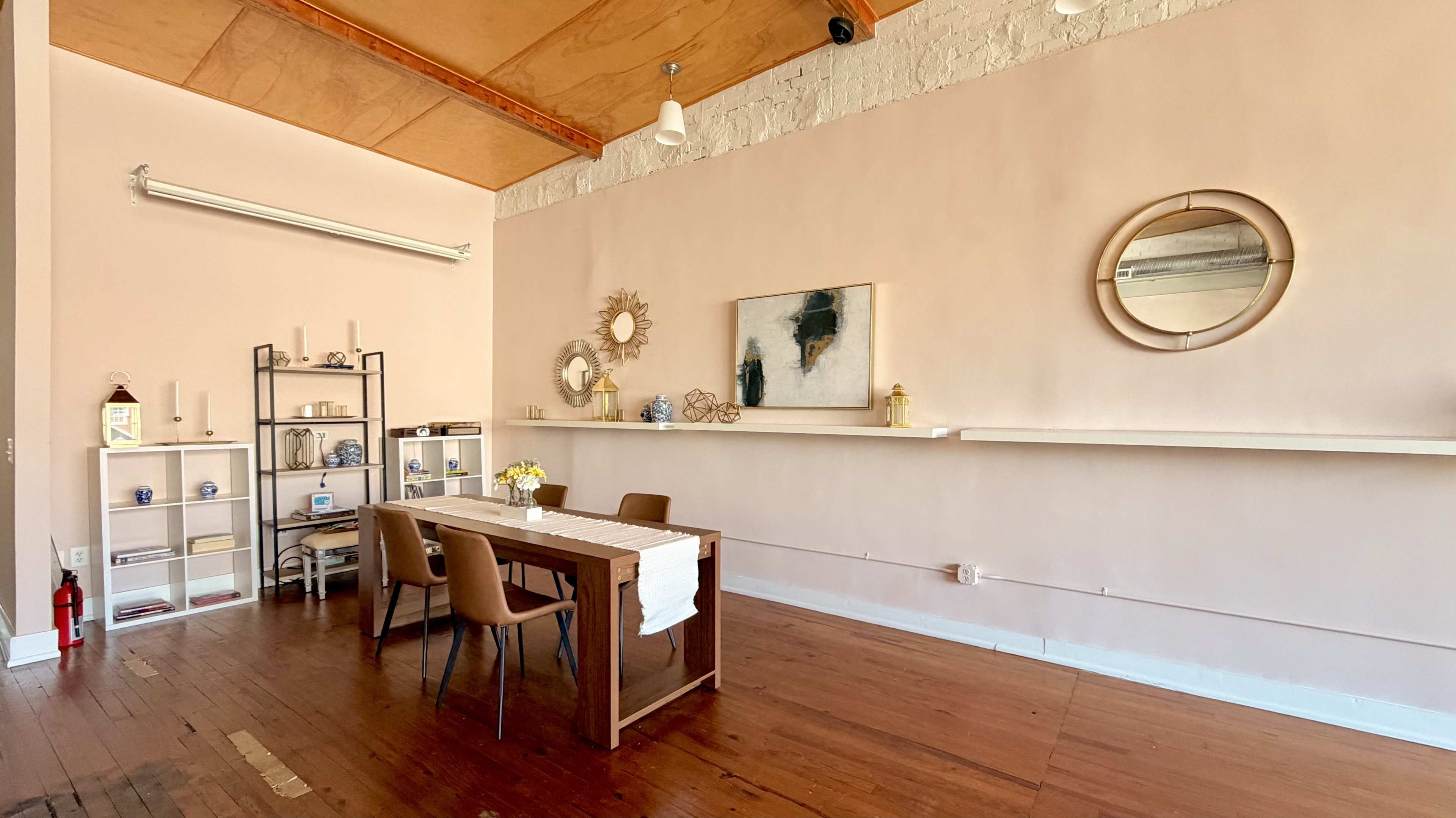 The room features a wooden dining table with four chairs, a decorative shelf on the left, mirrors on the wall, and light-colored walls with a wooden ceiling.
