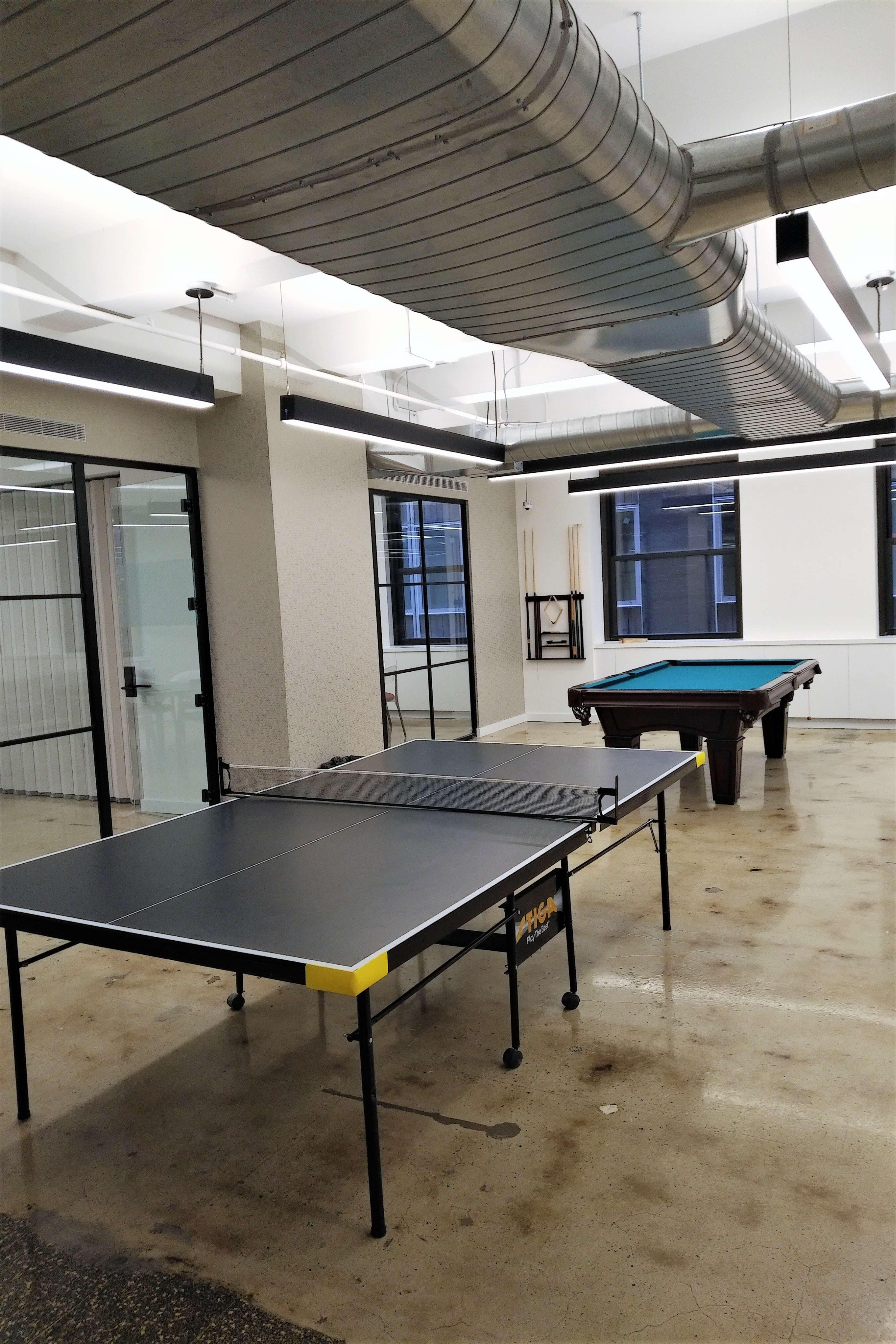 The image shows a room with a ping pong table and a billiards table, featuring industrial-style ventilation pipes above.