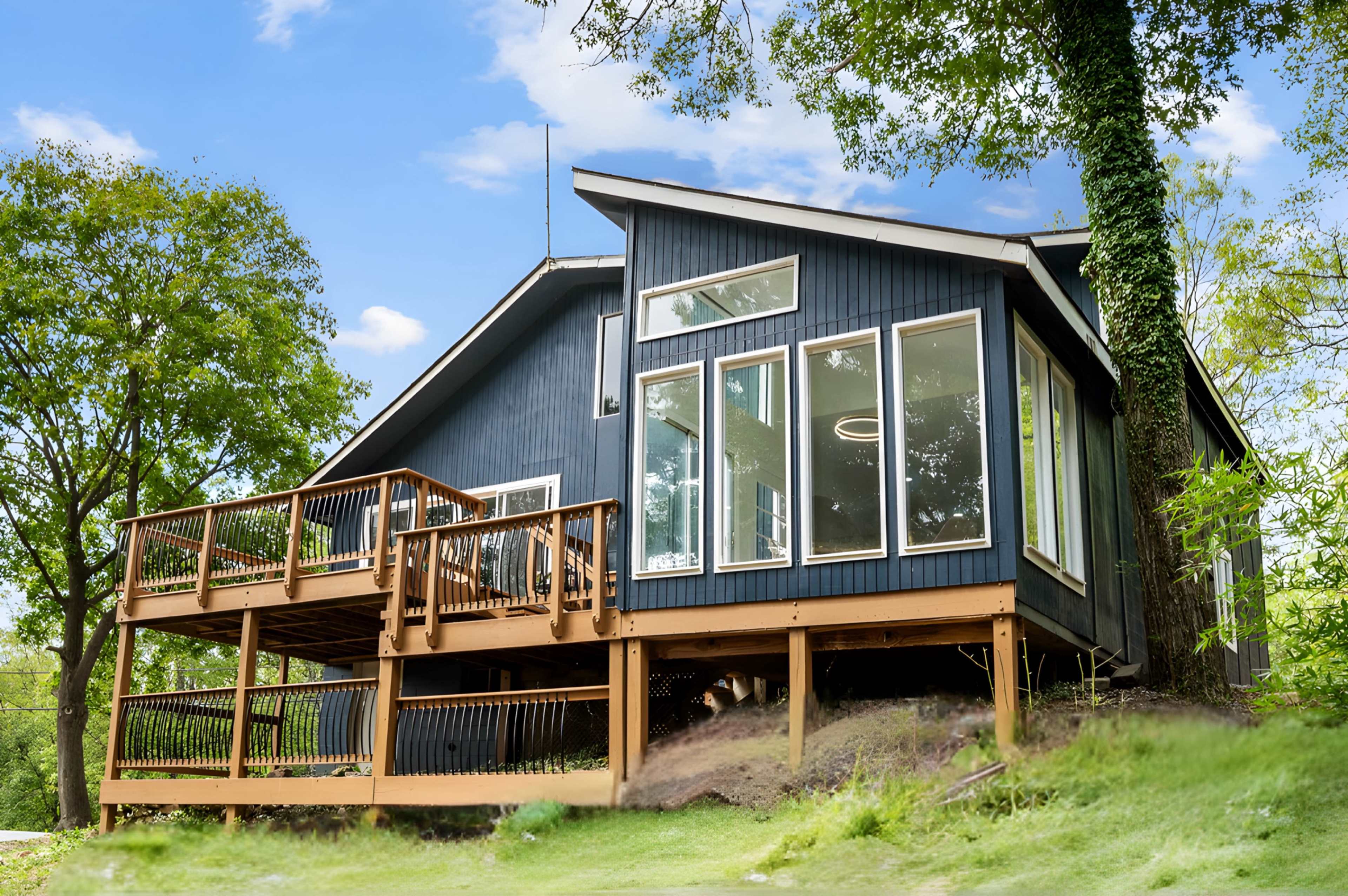 A modern house with large windows and a wooden deck is situated on a grassy slope, surrounded by trees.