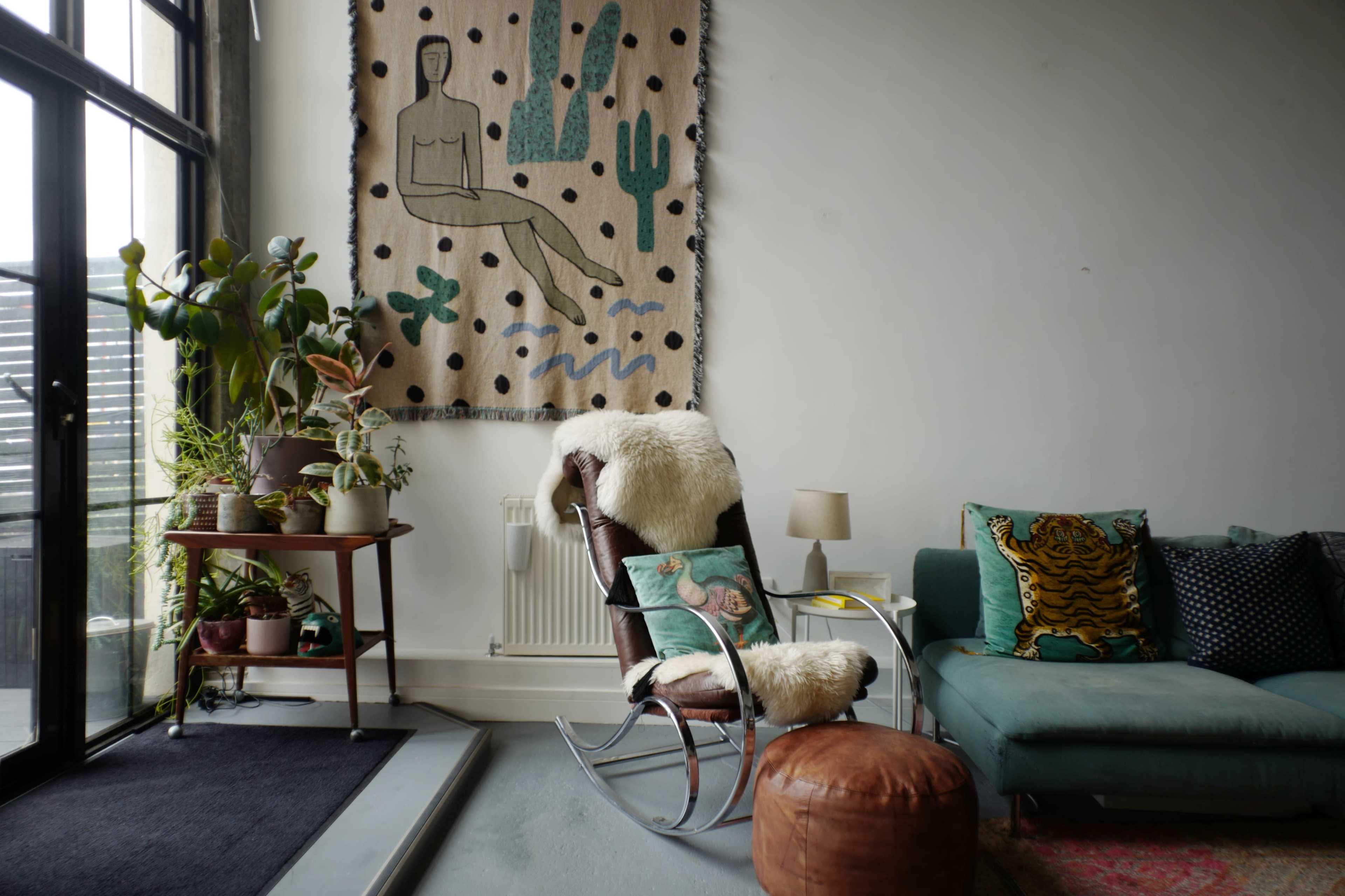 A modern living room features a rocking chair with a cozy throw, a green sofa adorned with decorative pillows, and a large wall tapestry depicting a figure surrounded by plants.