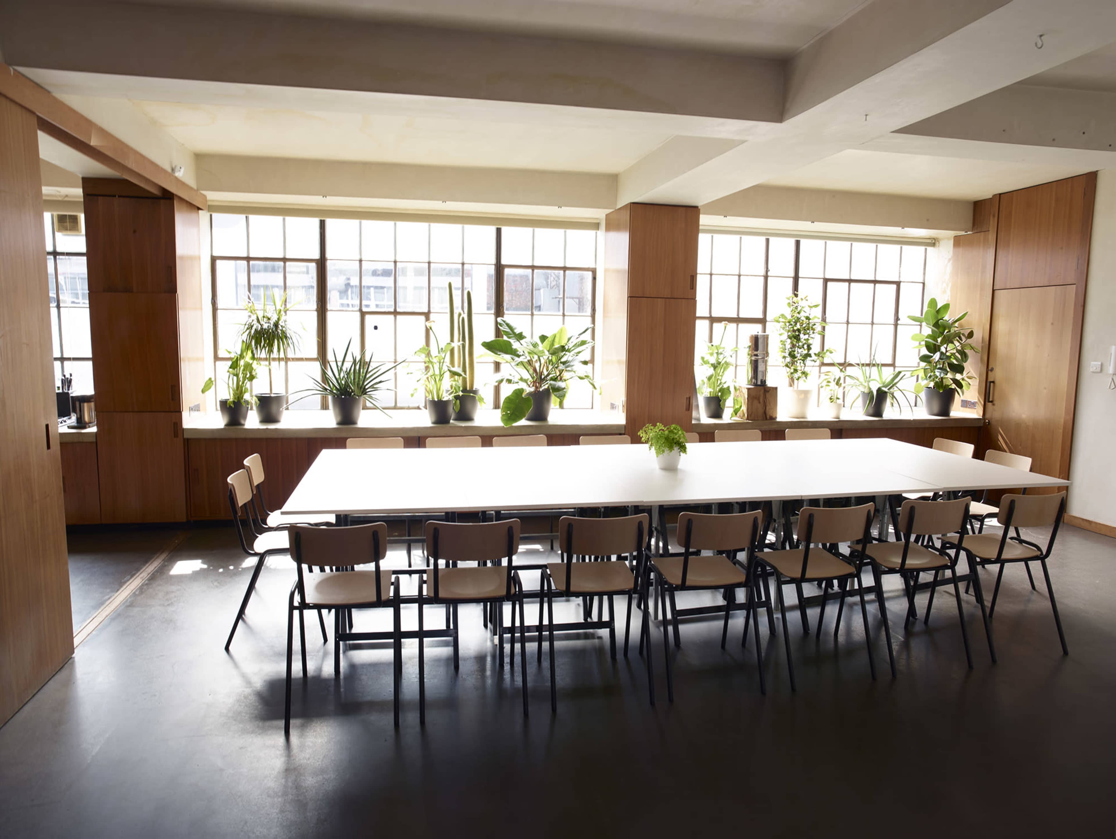 A long table with chairs is set in a well-lit conference room, featuring large windows lined with potted plants.