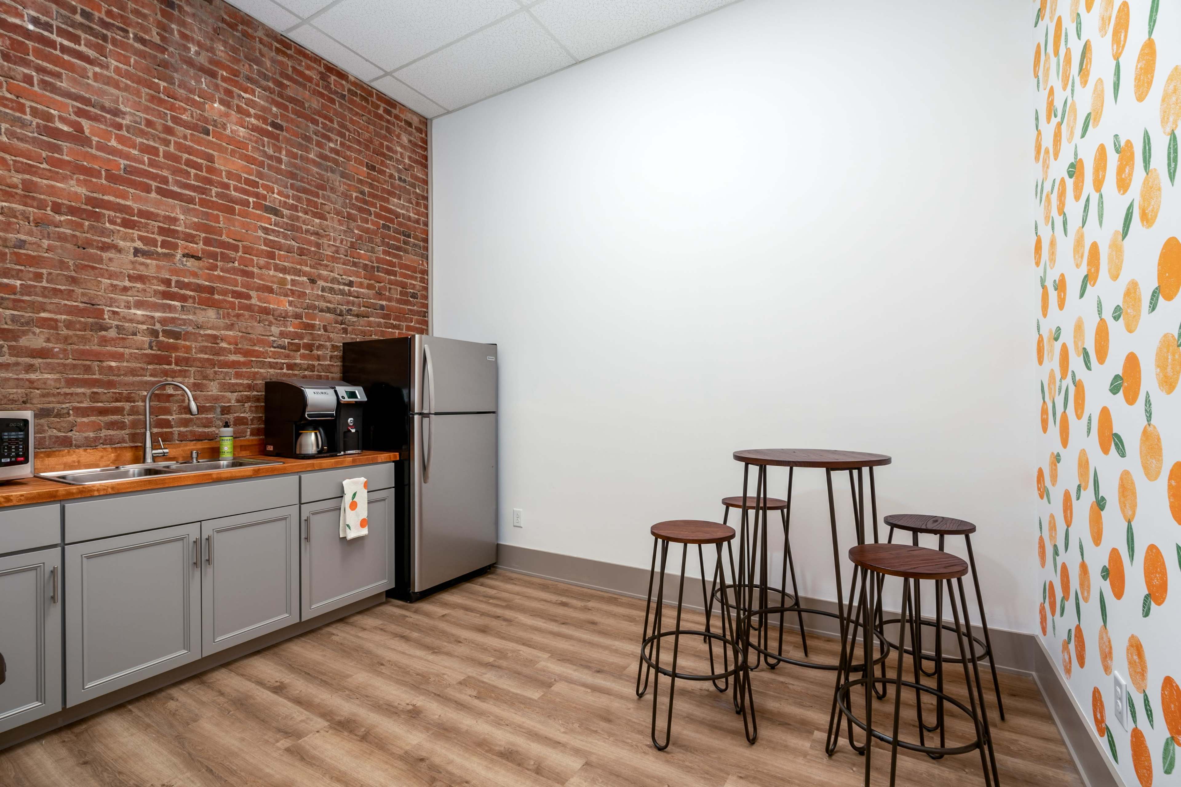 A minimalist kitchen area with exposed brick walls, a stainless steel refrigerator, a wooden countertop, and a small dining set featuring three bar stools.