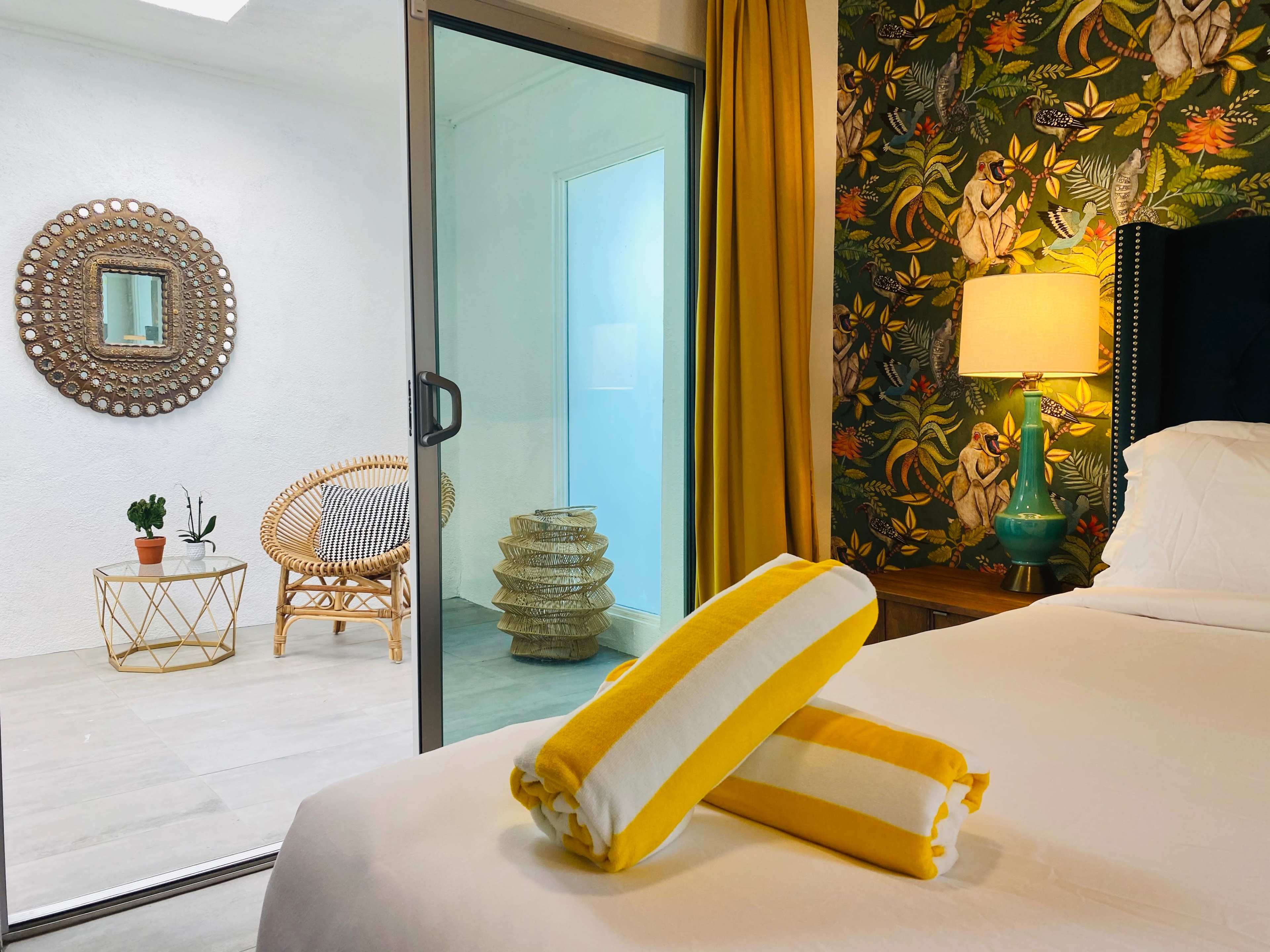 A bright bedroom features a bed with yellow-striped towels, a glass door leading to a brightly decorated sitting area with plants and a mirror.