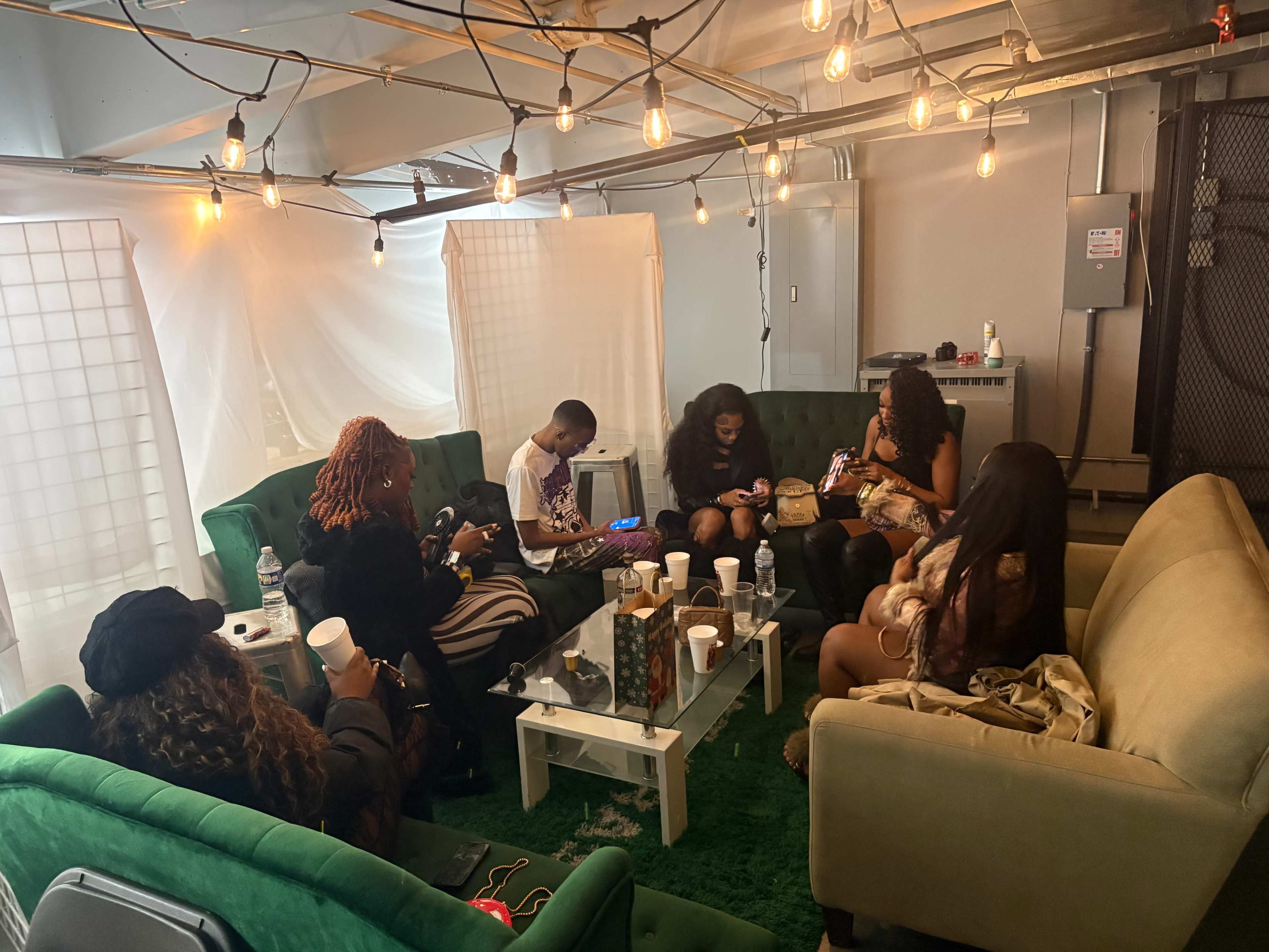 A group of people sits on green couches in a well-lit room, engaging with their phones and each other.