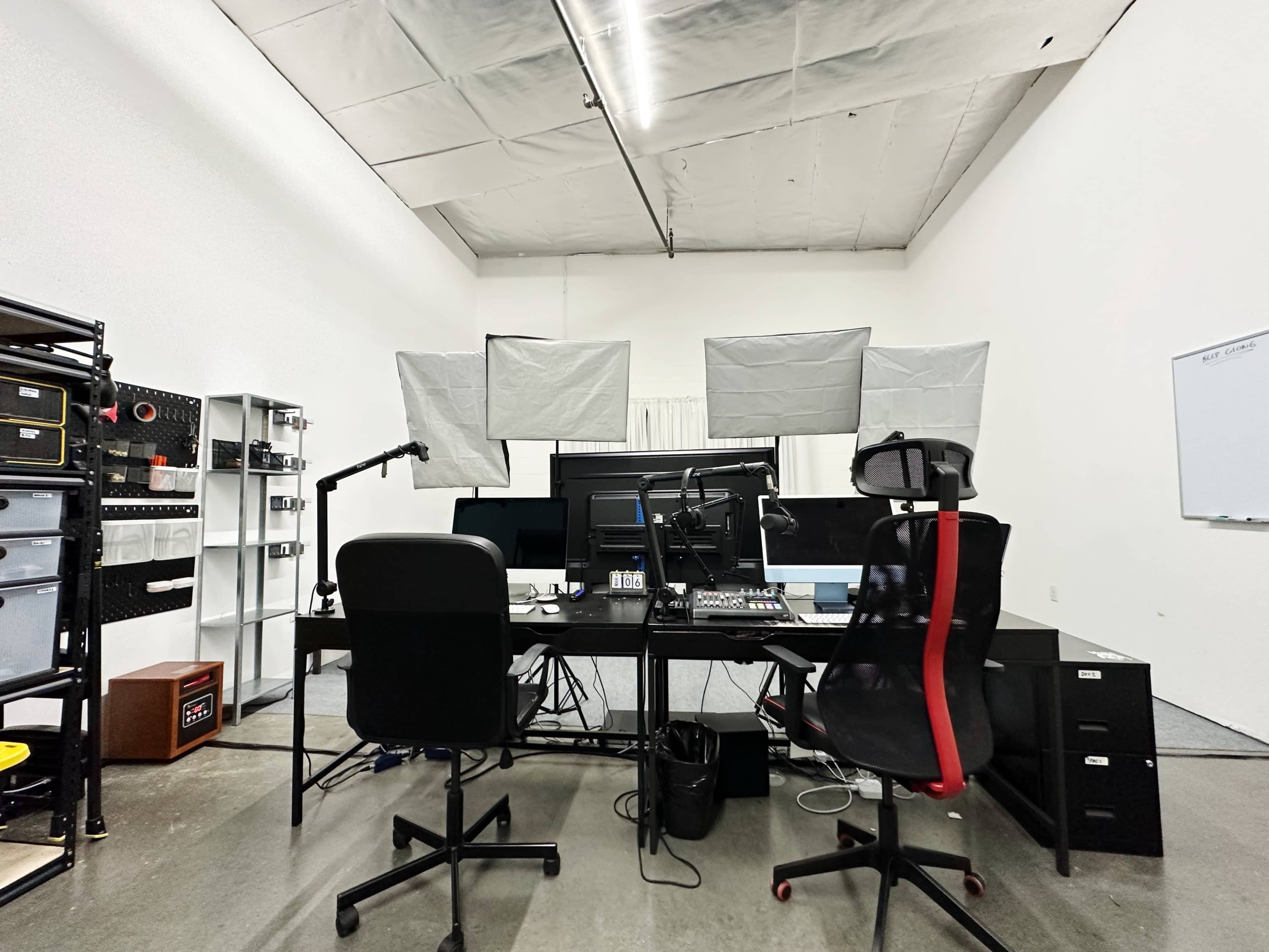 A workspace with a large black desk, two chairs, several computer monitors, and a microphone set up under softbox lighting.