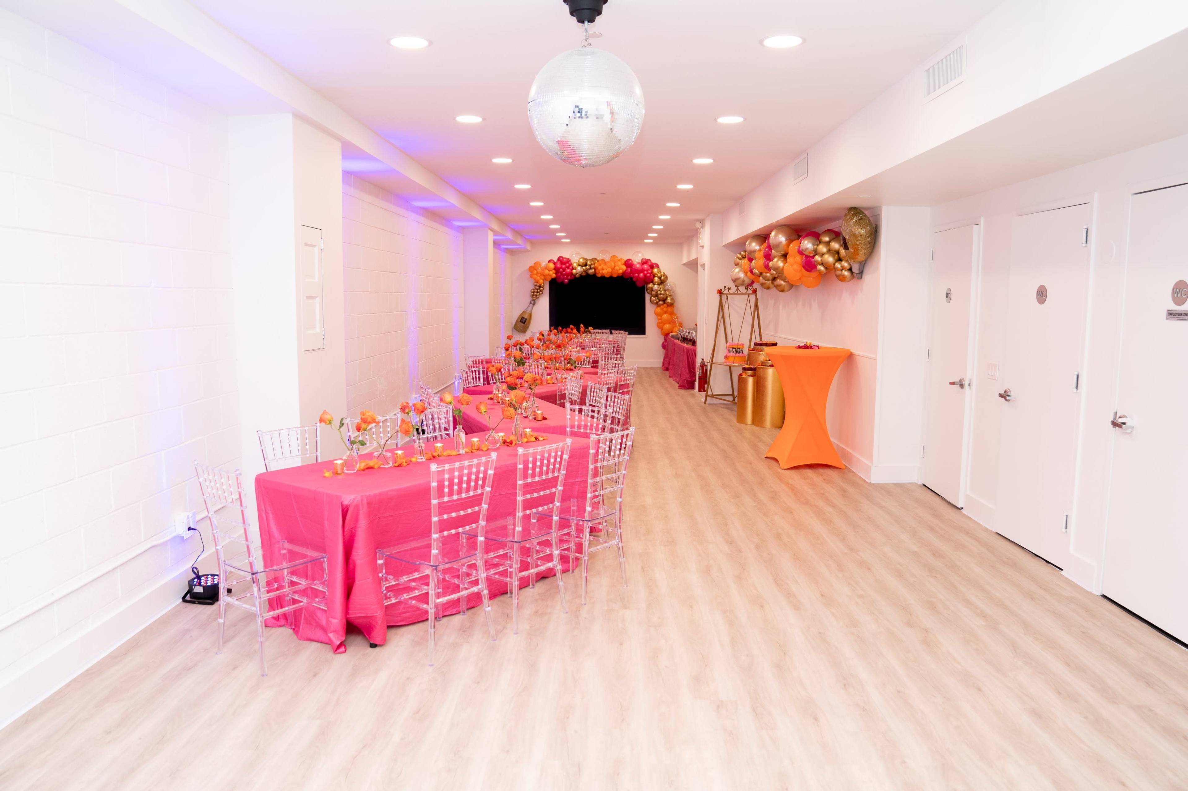 The image shows a decorated event space with a long table set with pink tablecloths, clear chairs, and colorful balloons in the background.
