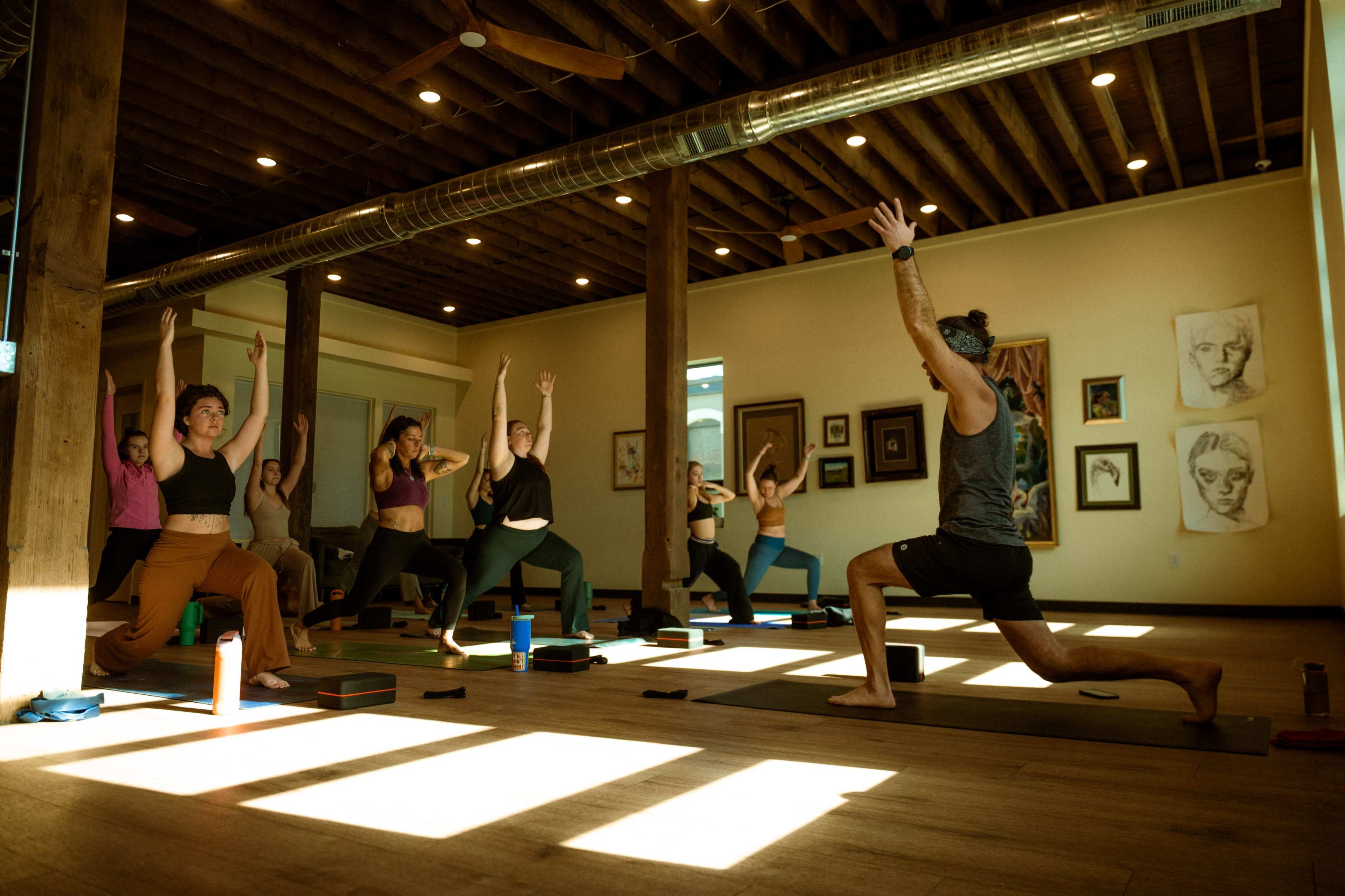 A group of individuals participates in a yoga class in a spacious studio with wooden beams and artwork on the walls.