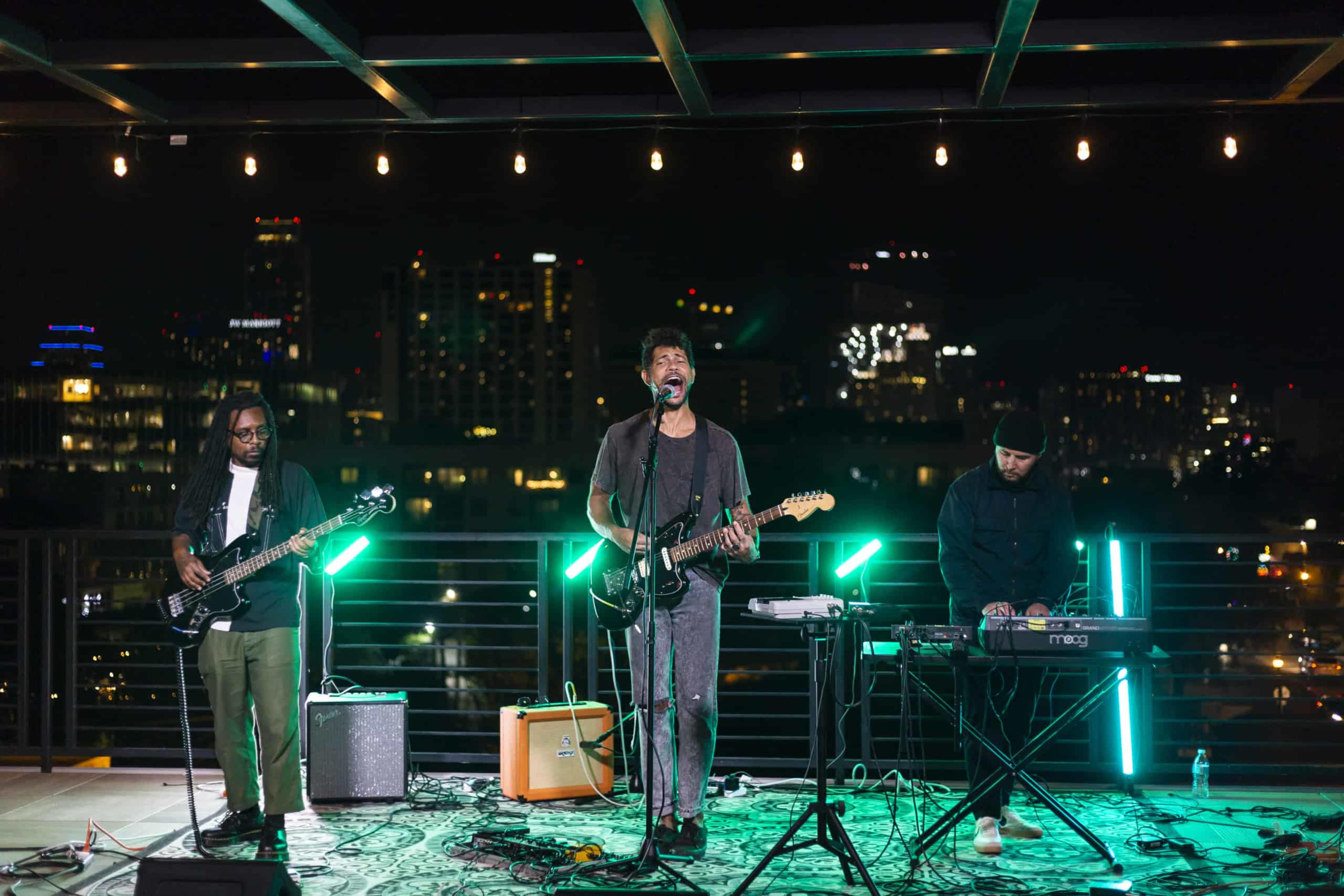 A band performs on a rooftop at night, with city lights visible in the background.