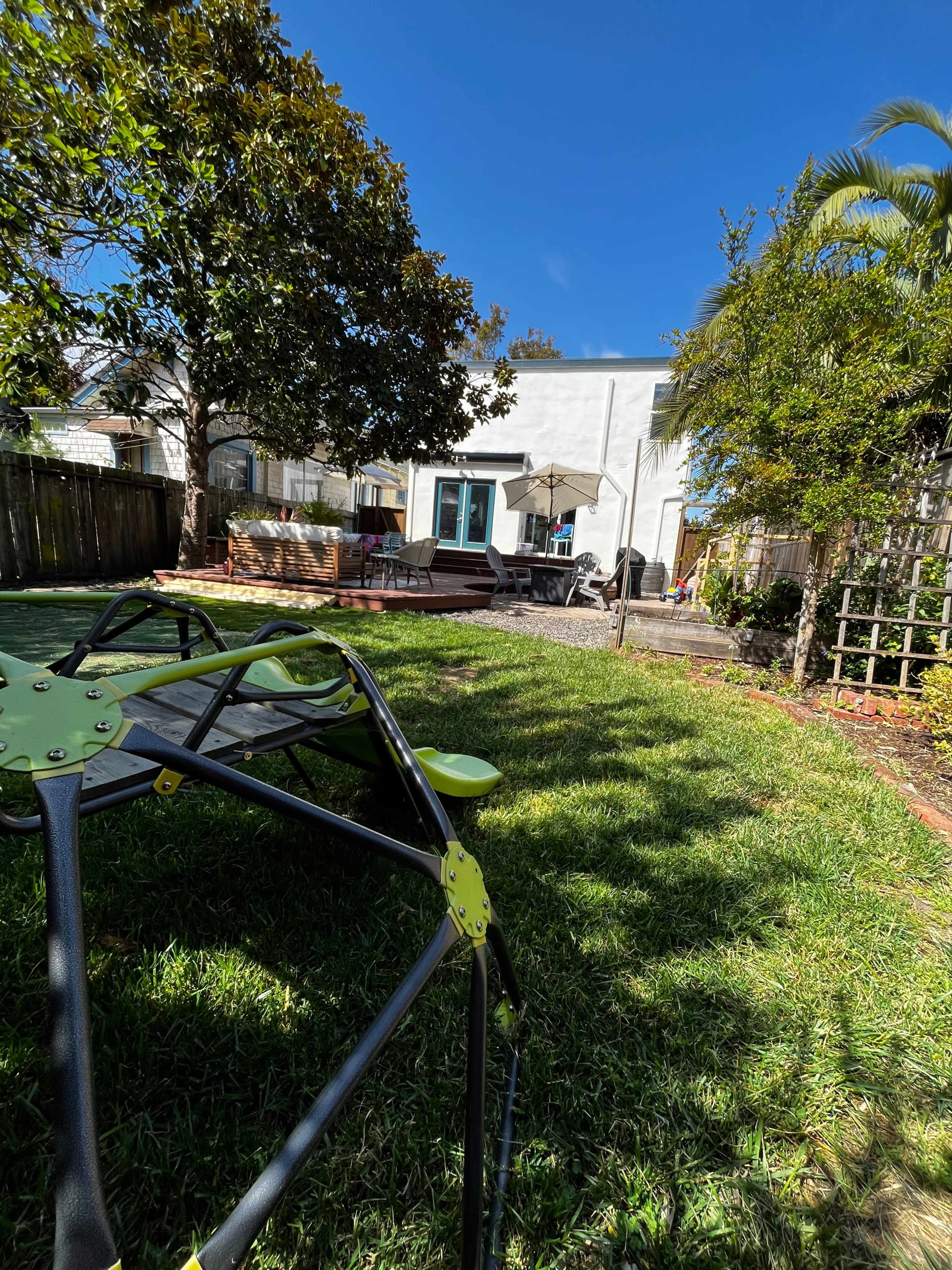 The photo shows a backyard scene featuring a green lawn, a patio with outdoor furniture, and a house in the background under a clear blue sky.