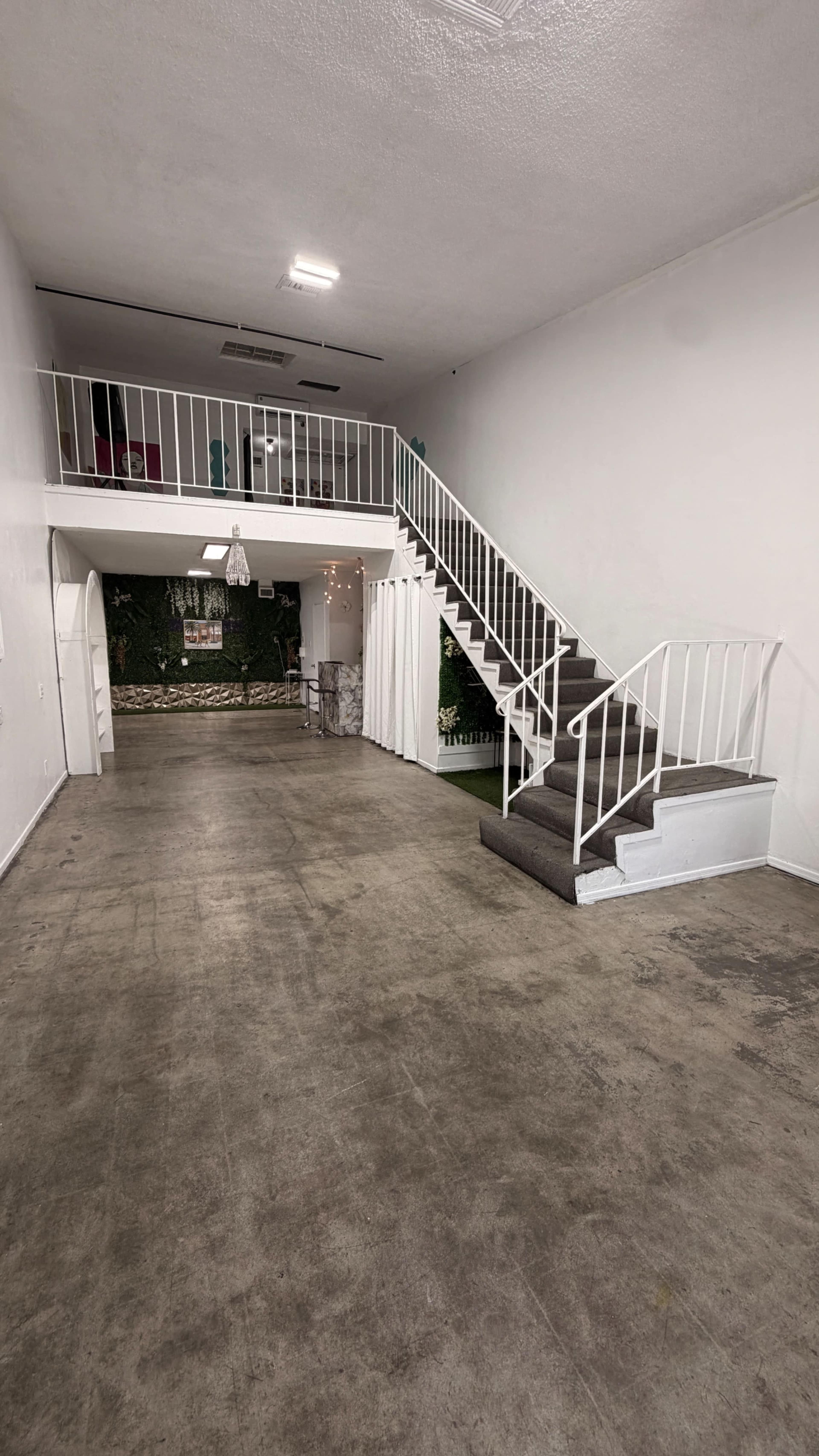 The image shows an empty interior space with a staircase leading to an upper level, featuring a concrete floor and a small green area at the back.