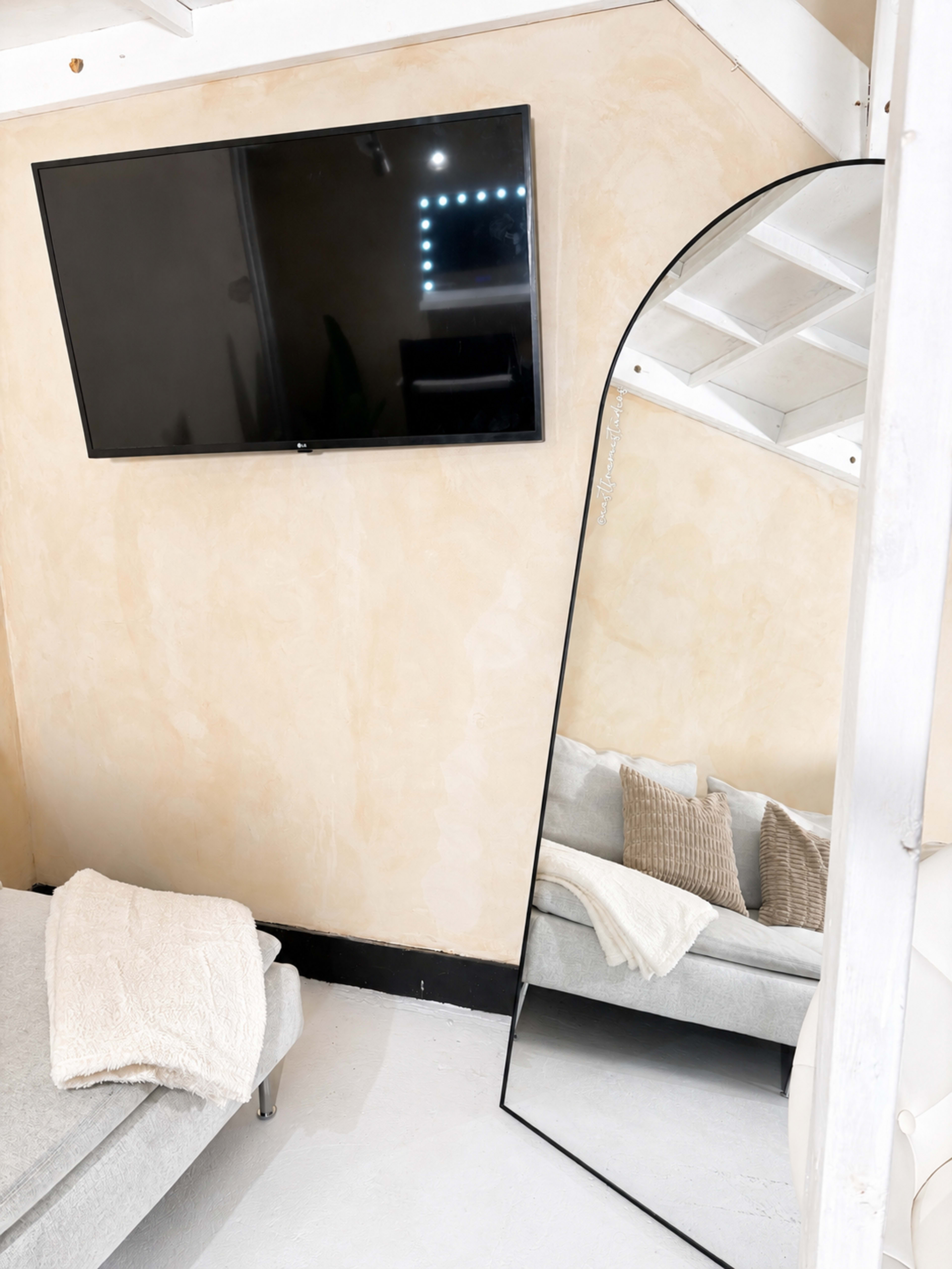 The image shows a minimalistic room featuring a television mounted on the wall, a large arched mirror, and a gray couch with decorative pillows.