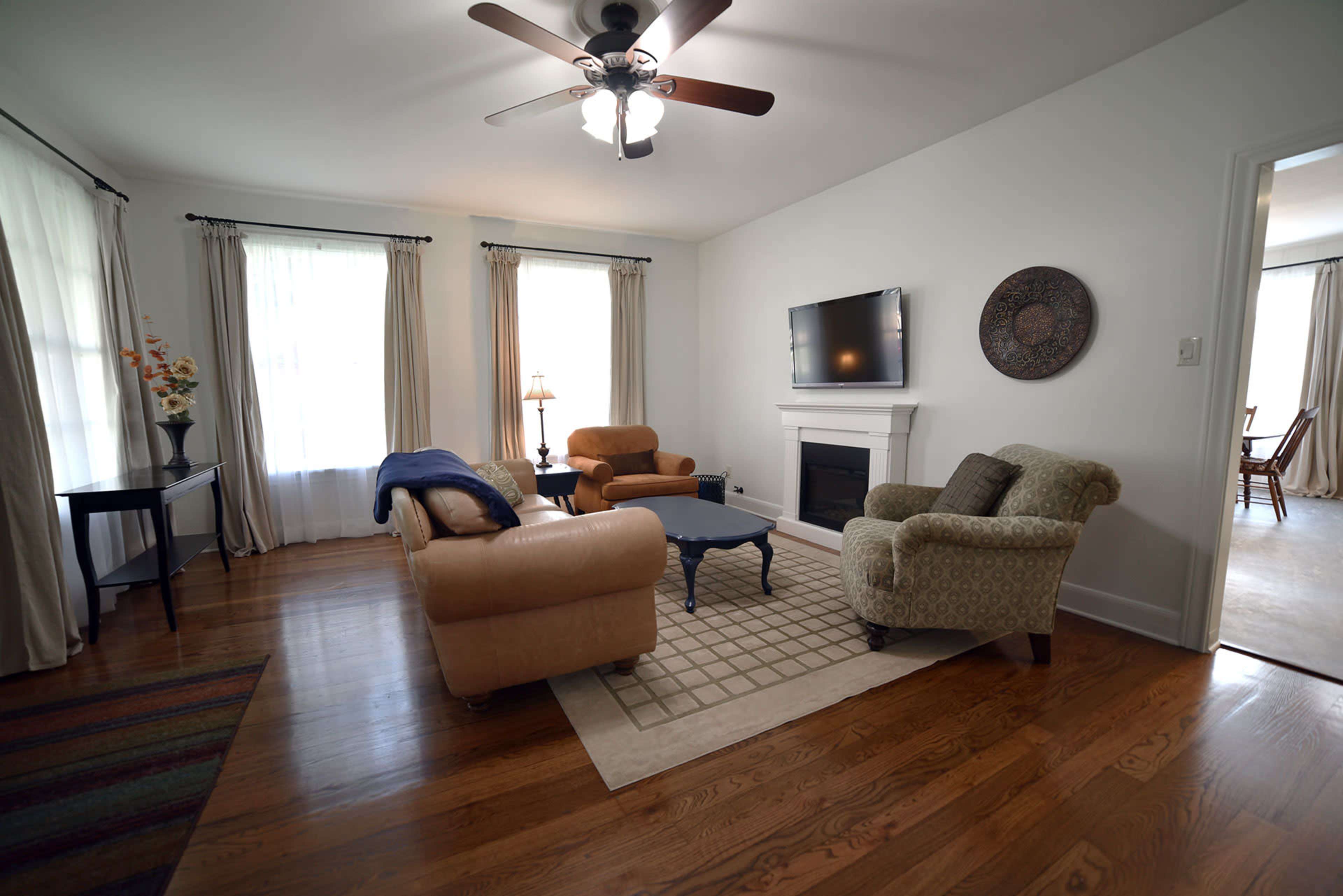 A living room with a ceiling fan, a sofa, two armchairs, a coffee table, a fireplace, and large windows dressed with curtains.