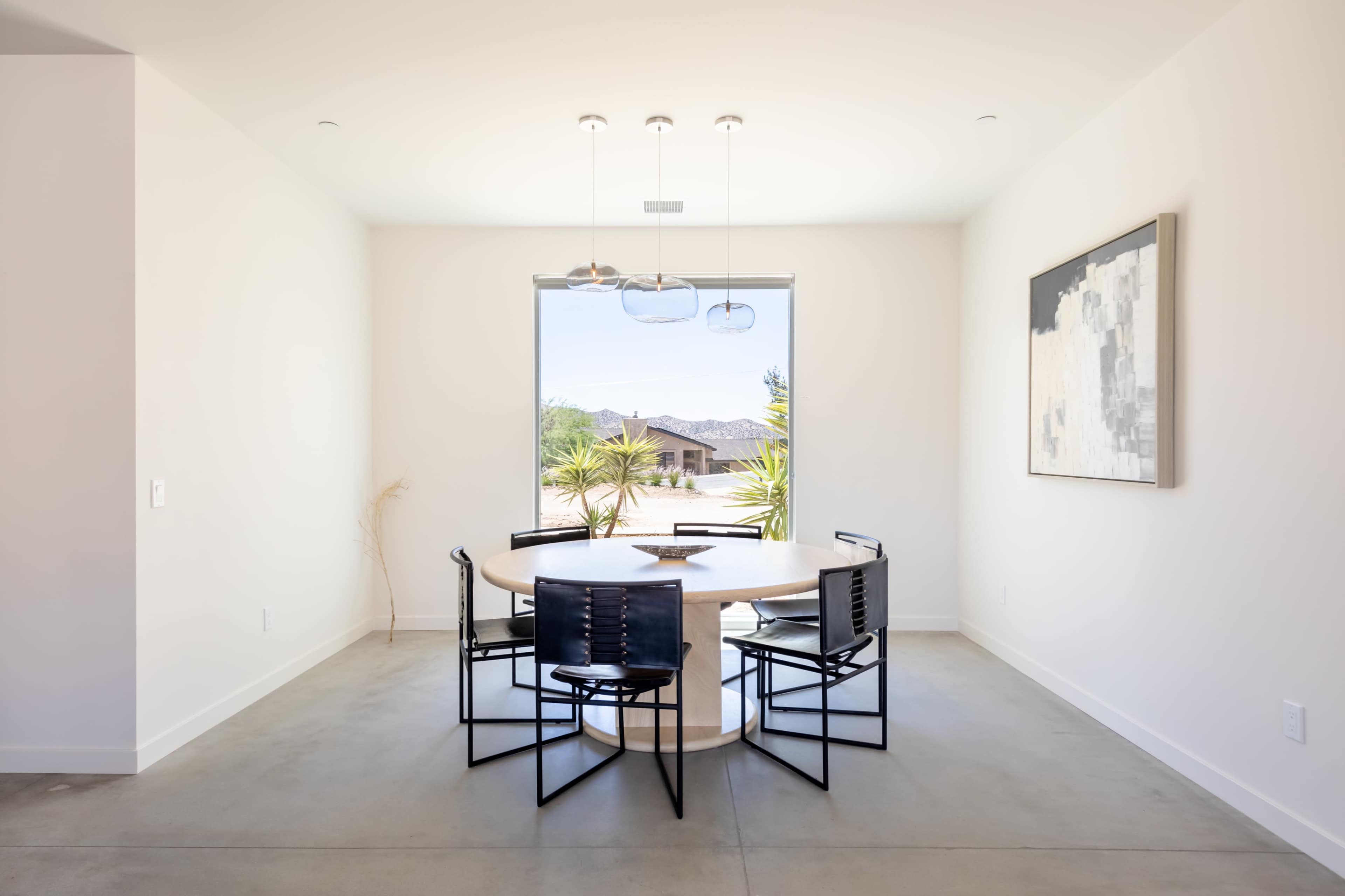 A round dining table surrounded by six black chairs is positioned in a minimalist room with large windows showcasing outdoor greenery.