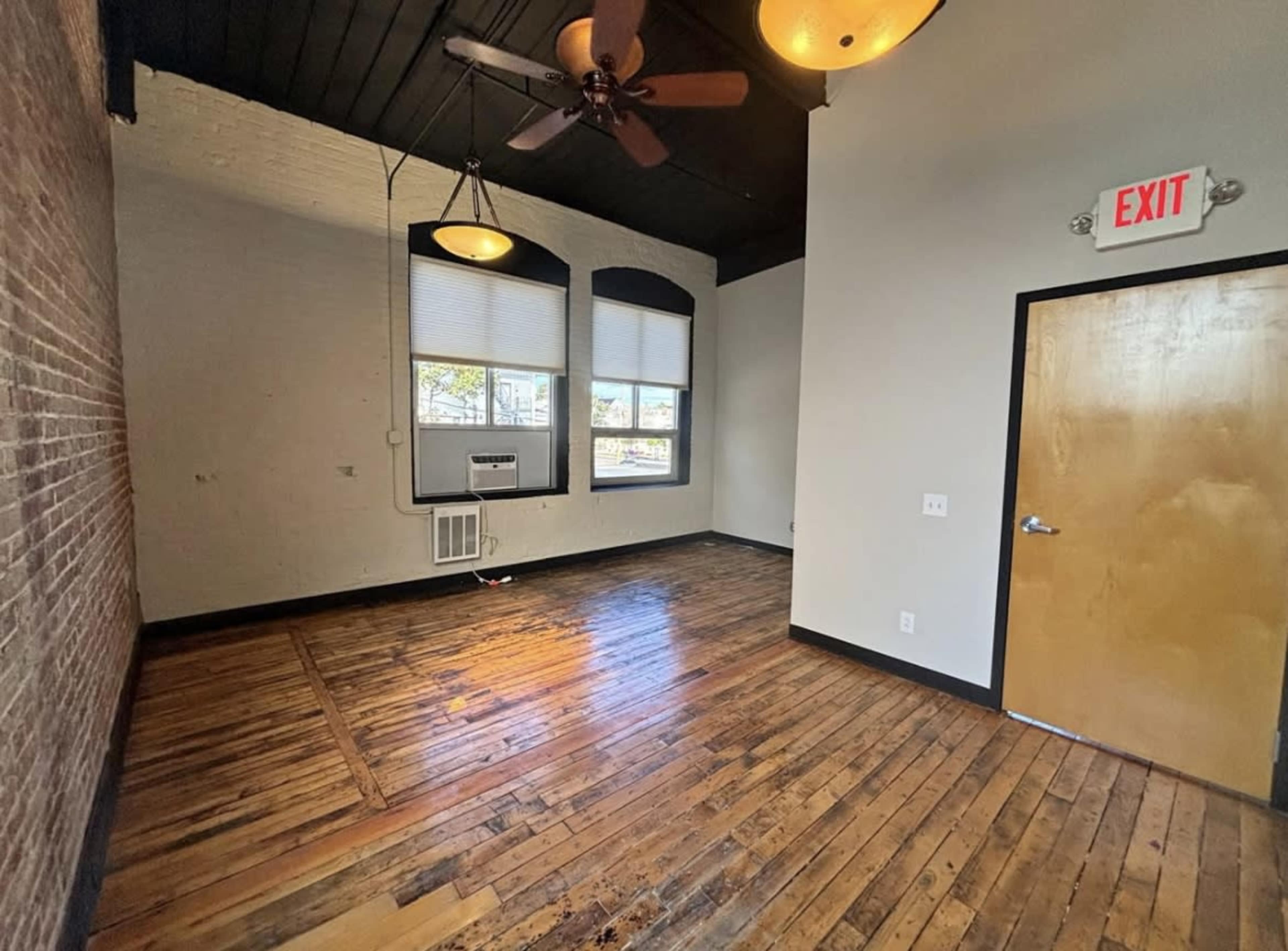 The image shows an empty room with wooden floors, a ceiling fan, air conditioning unit, and exposed brick walls.