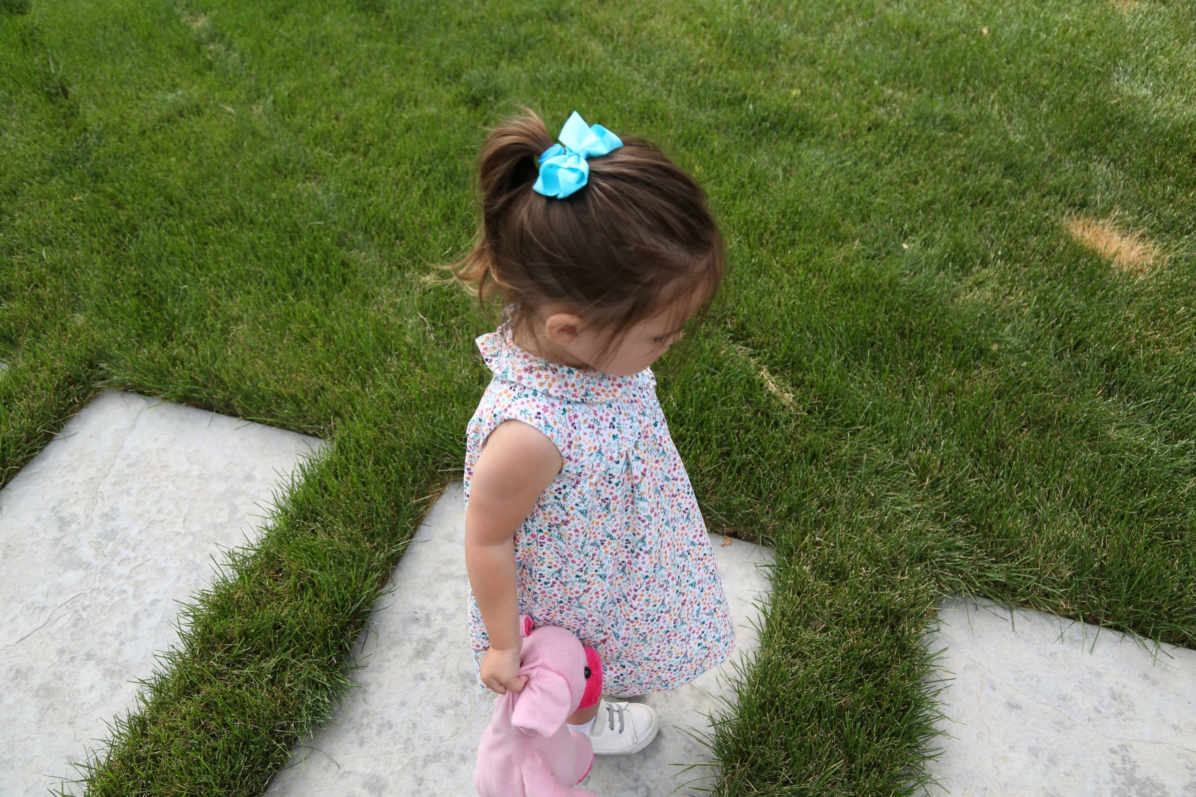 A young child in a floral dress holds a pink stuffed animal while walking along a grassy pathway.